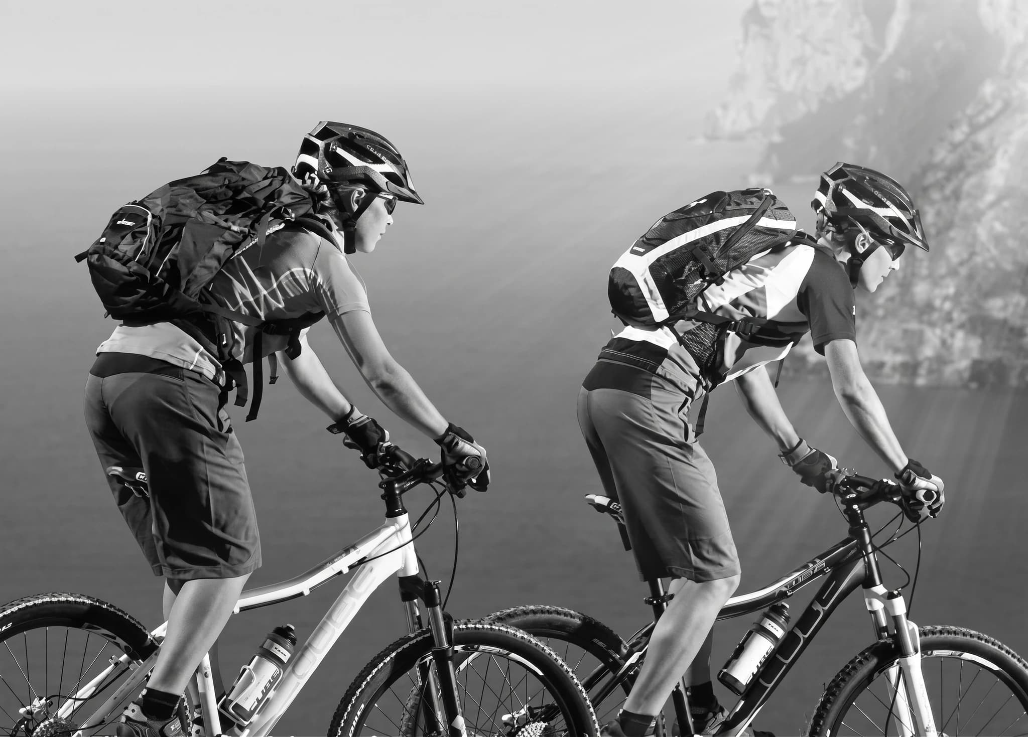 Två cyklister på mountainbikes med ryggsäckar, cyklar i landskap med berg och vatten.