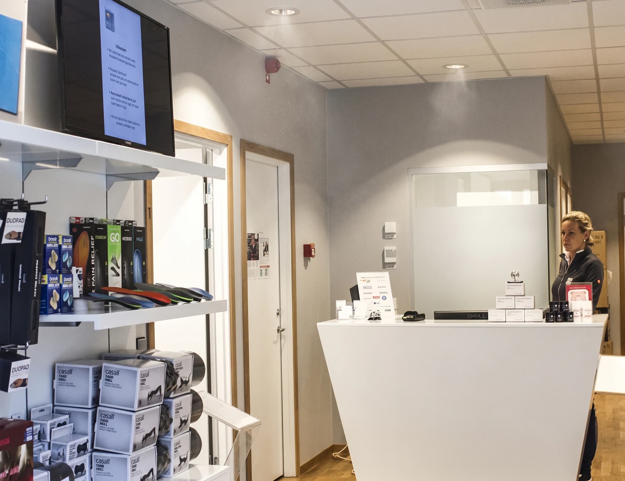 En kvinna står bakom en vit receptionsdisk i en klinik, med hyllor med produkter till vänster och en dörr till höger.