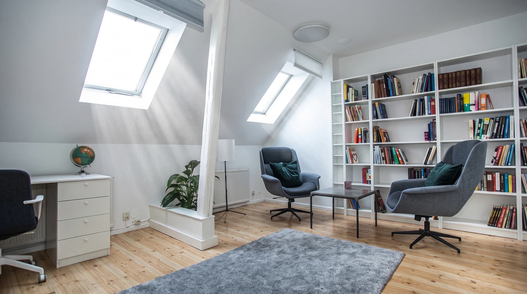 Ett ljust och modernt terapirum med takfönster, bokhyllor fyllda med böcker, två gråa fåtöljer, ett soffbord och en grå matta på trägolv.