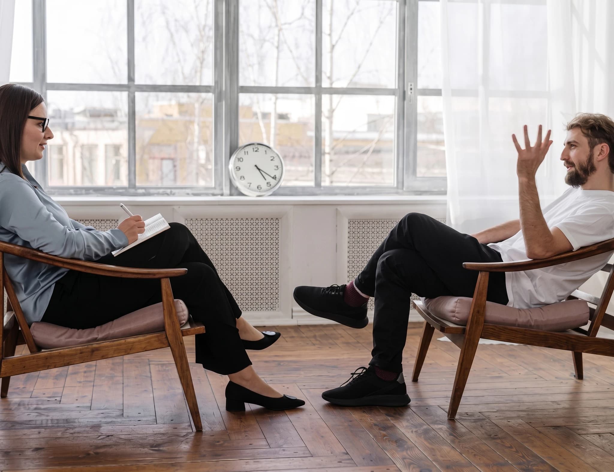Terapeut med glasögon sitter och skriver i en anteckningsbok medan en patient sitter mittemot och pratar.