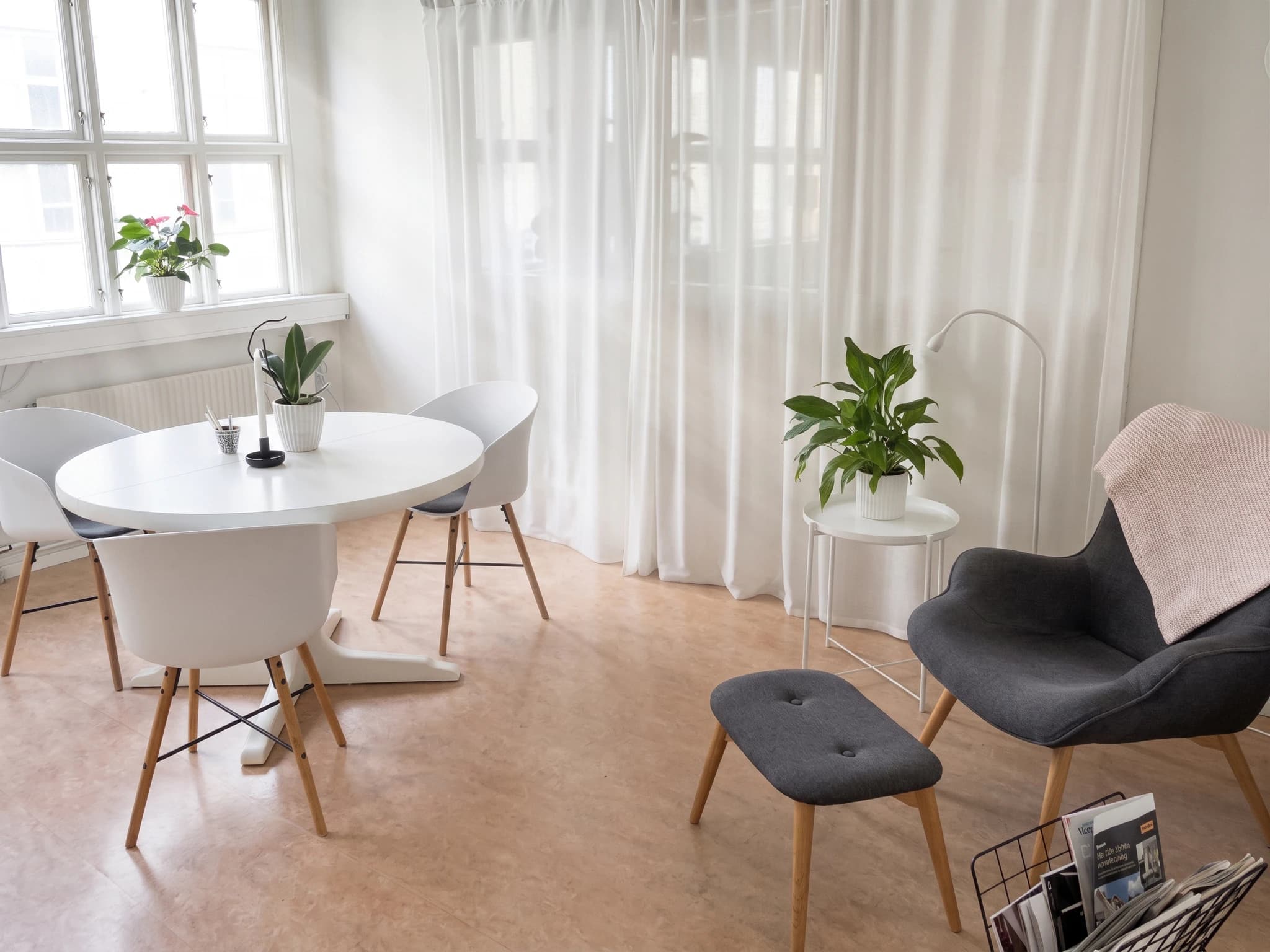 Ljus och modern väntrumsmiljö med ett runt vitt bord, vita stolar, en mörkgrå fåtölj med fotpall, vita gardiner och växter.