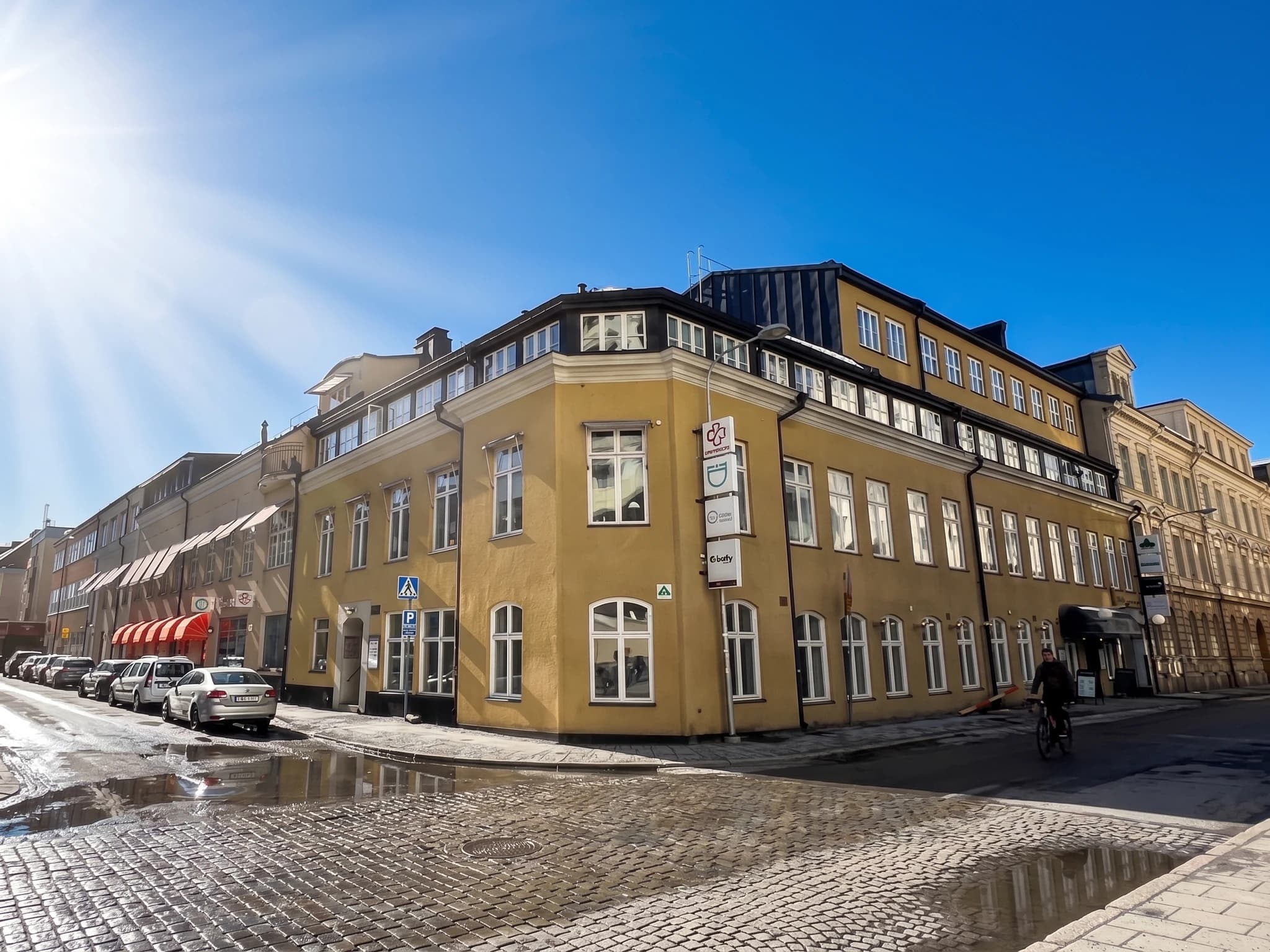 Gul byggnad i ett hörn med bilar parkerade längs gatan och en person som cyklar på en solig dag.