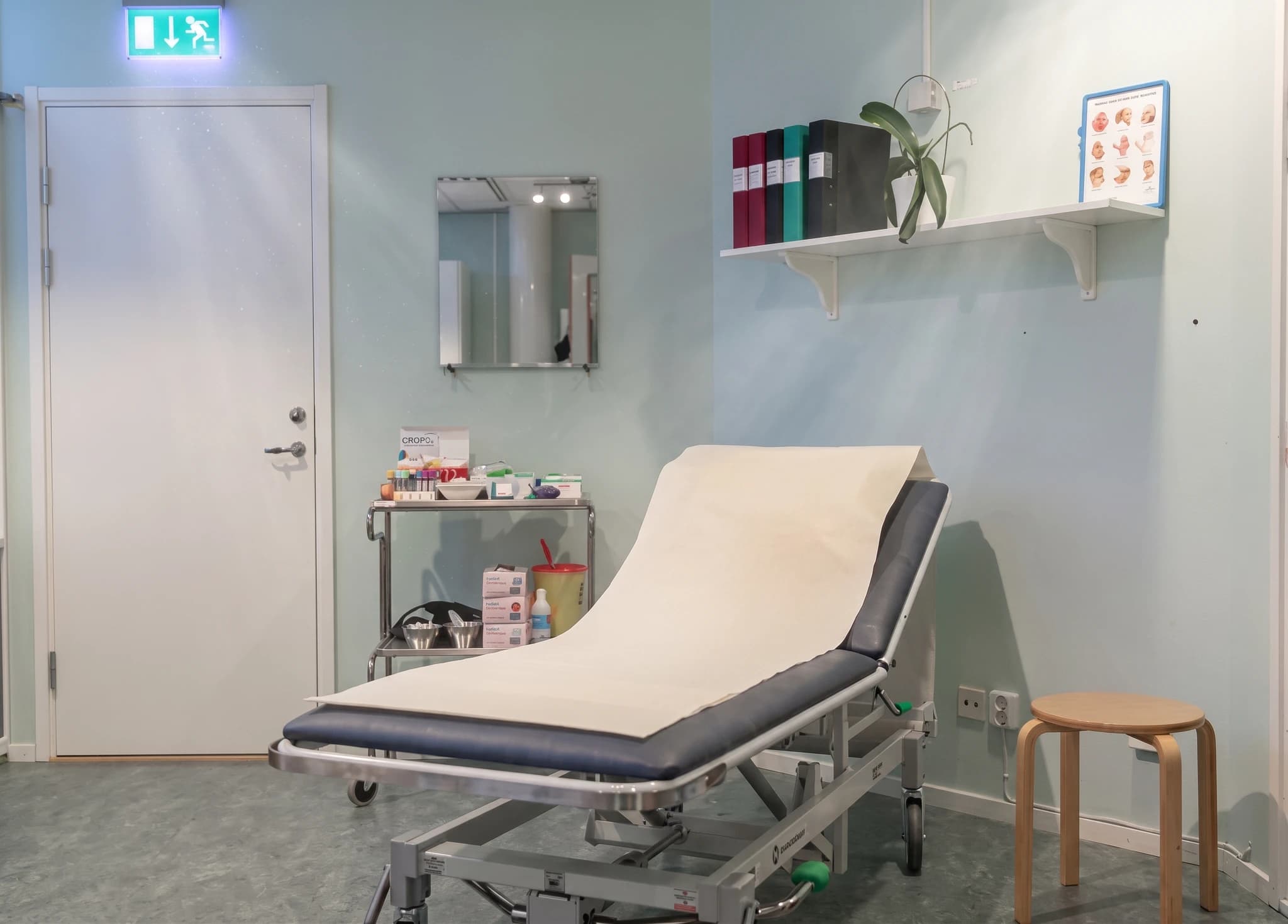 Ett undersökningsbord med vitt papper täckt, placerat i ett ljust kliniskt rum med medicinska förnödenheter på ett rullbord till vänster och en avföring till höger.