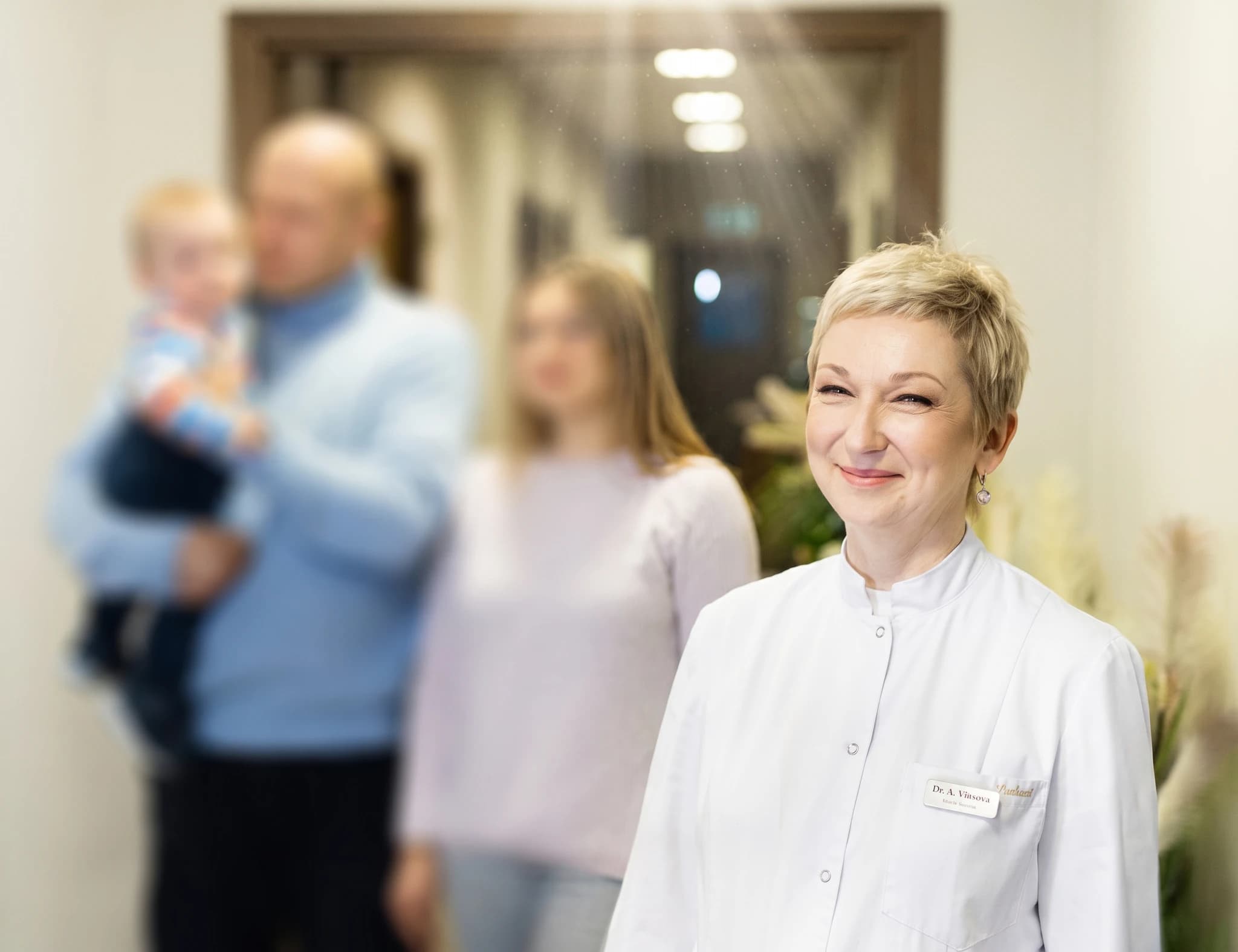 En leende läkare med kort blont hår bär en vit rock och står i en sjukhuskorridor. I bakgrunden skymtar en familj med ett barn.