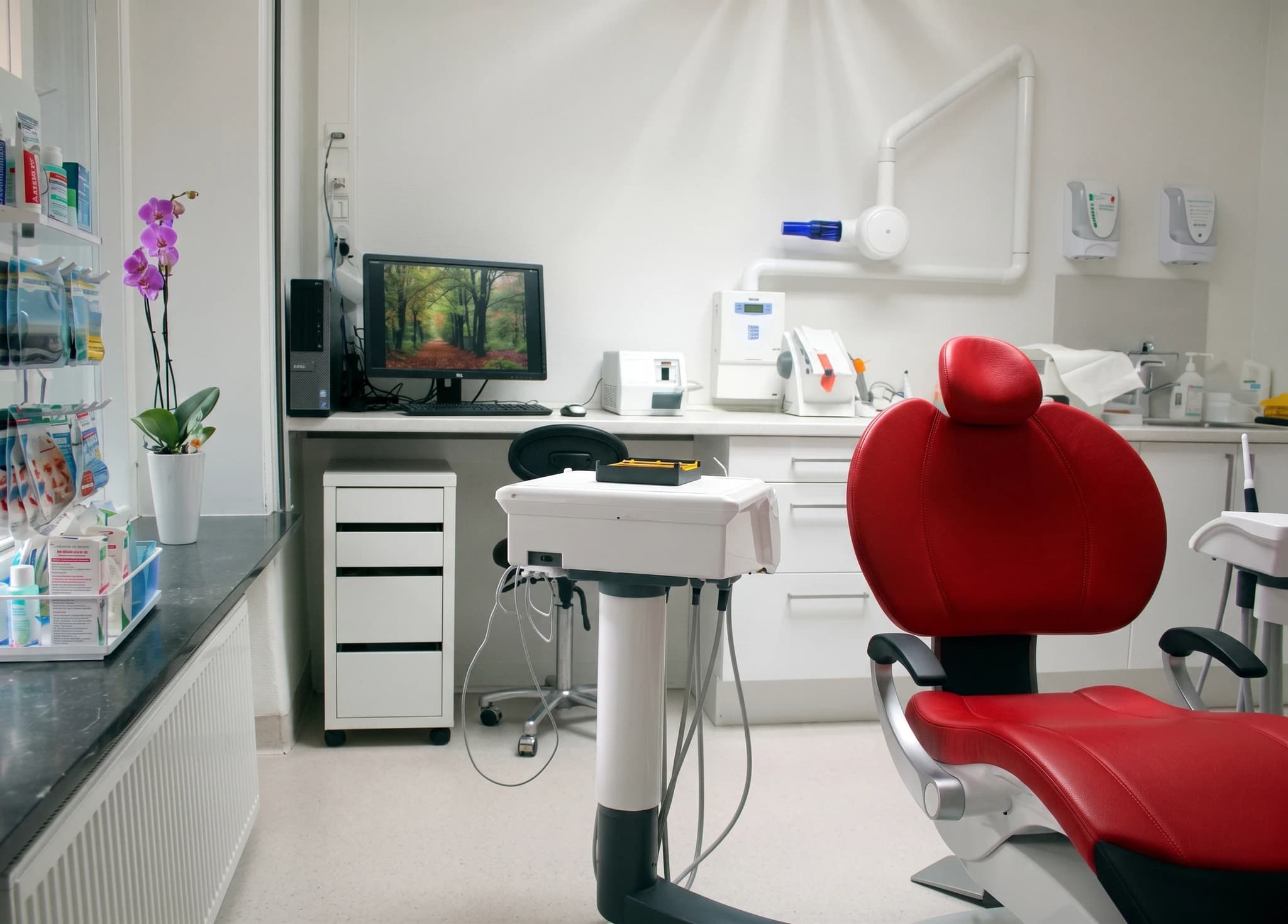 En modern tandläkarmottagning med en röd tandläkarstol, ett skrivbord med dator och medicinsk utrustning.
