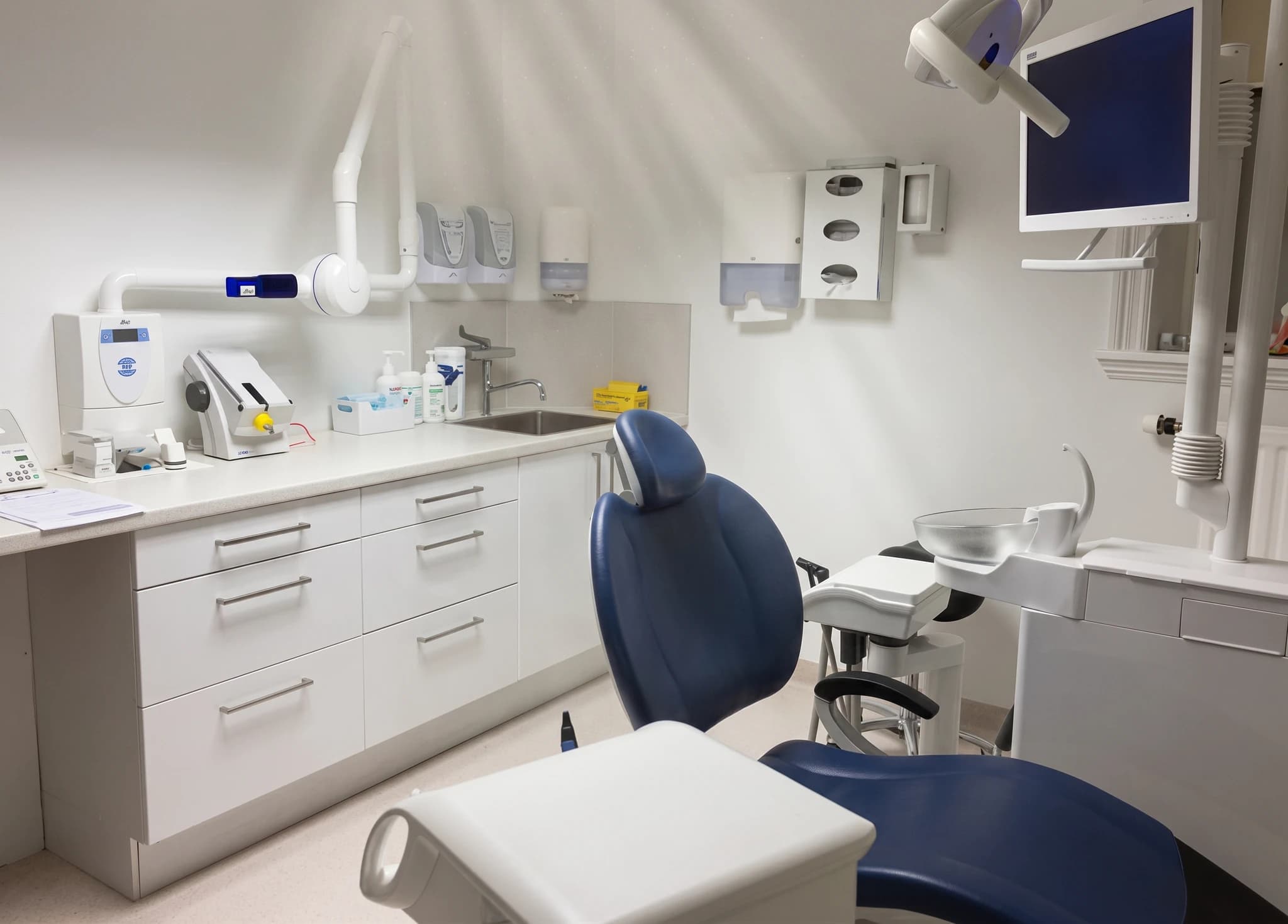 Ett modernt tandläkarrum med en blå och vit tandläkarstol, ett handfat och medicinsk utrustning monterad på väggen.