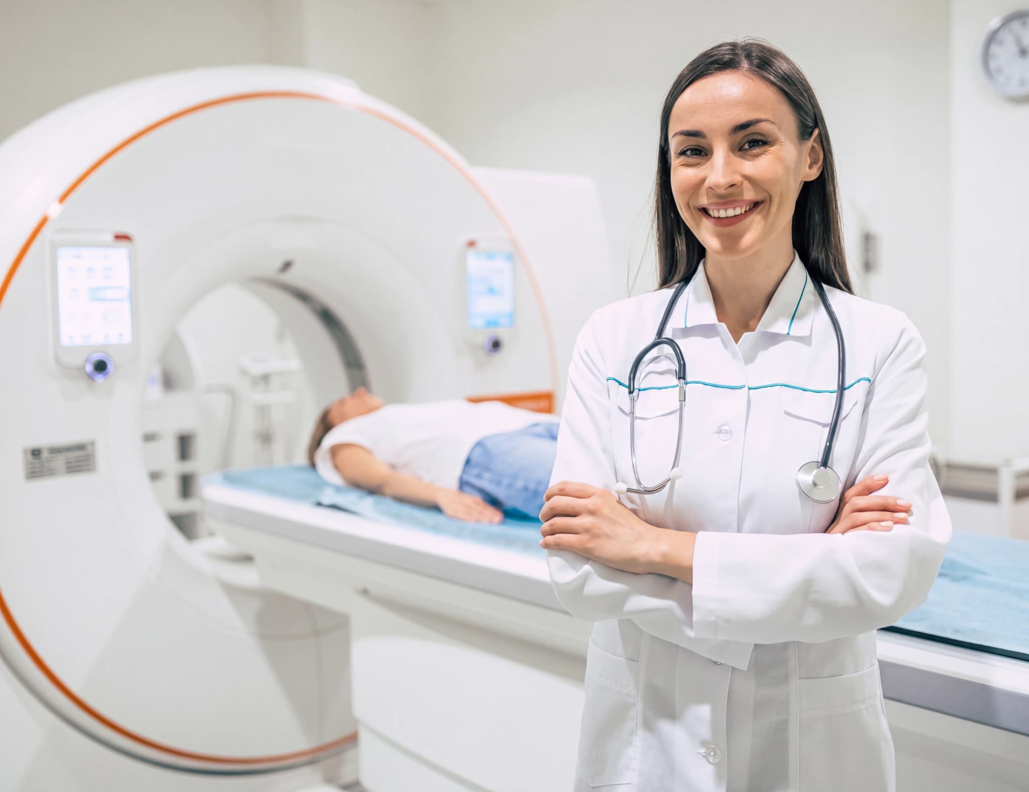 En leende läkare i vit rock med stetoskop står framför en CT-scanner där en patient ligger på britsen.