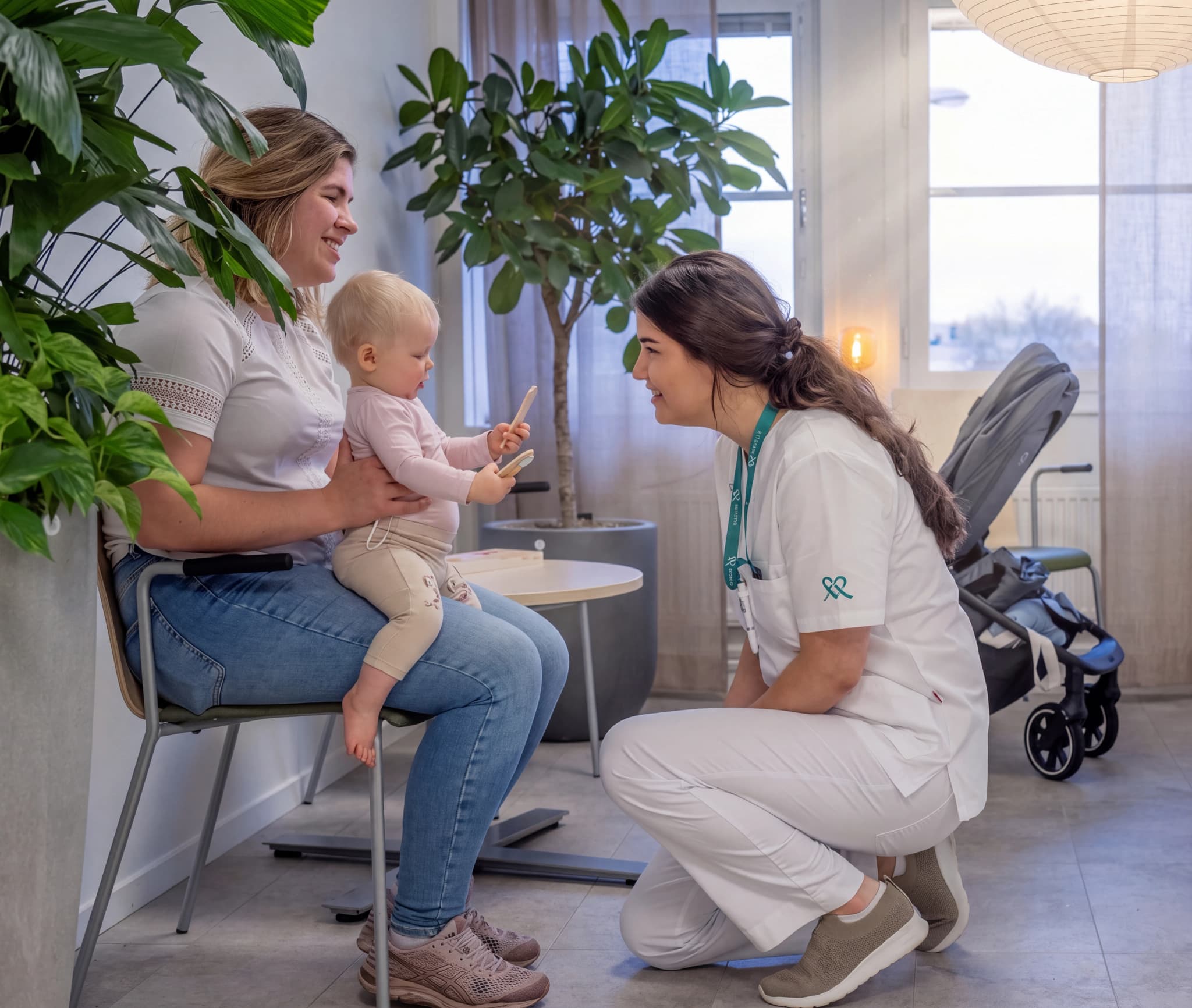 Sjuksköterska som undersöker ett spädbarn som sitter i sin mammas knä, med växter och en barnvagn i bakgrunden.
