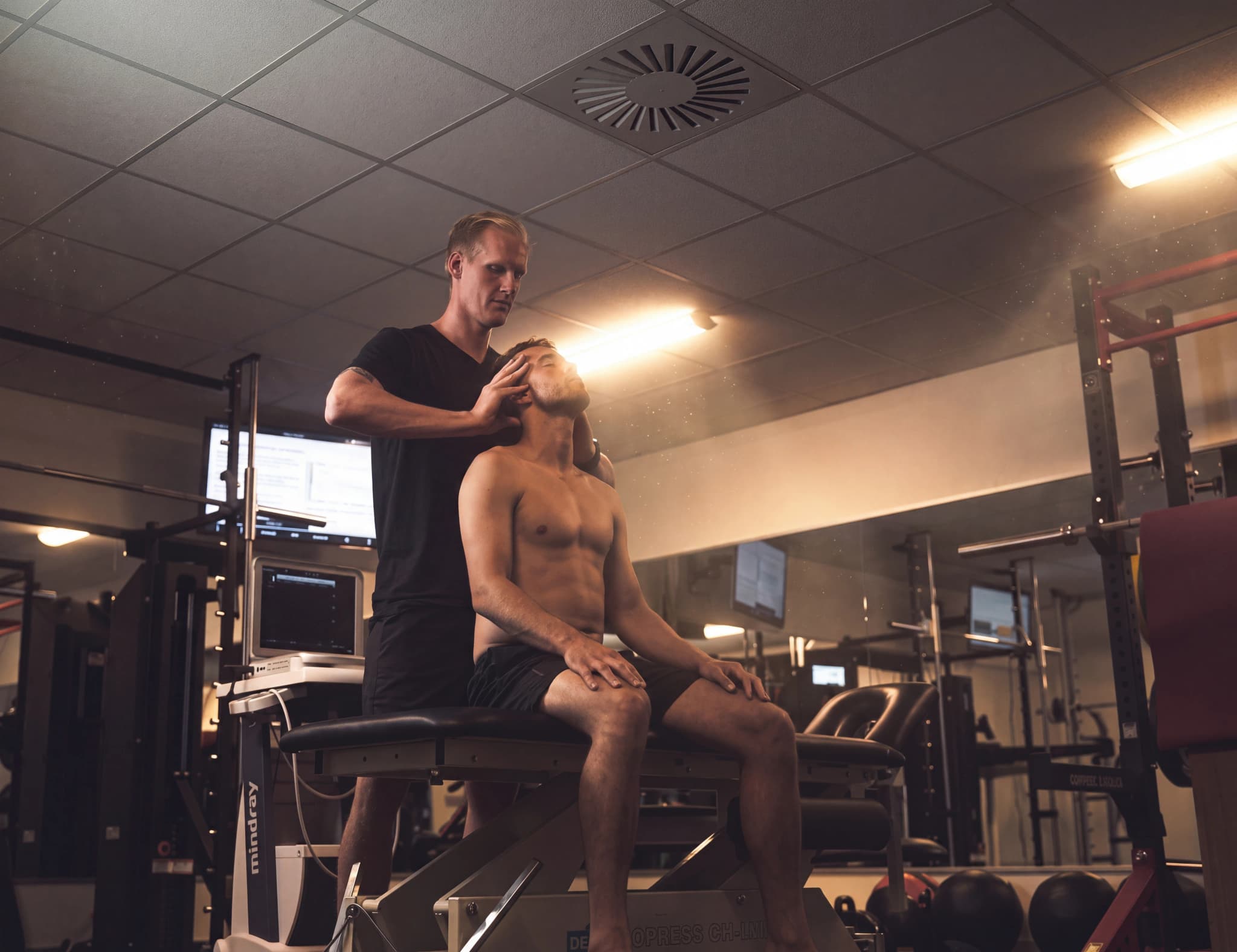 En fysioterapeut utför en nackjustering på en manlig idrottare i ett gym, med medicinsk utrustning synlig i bakgrunden.