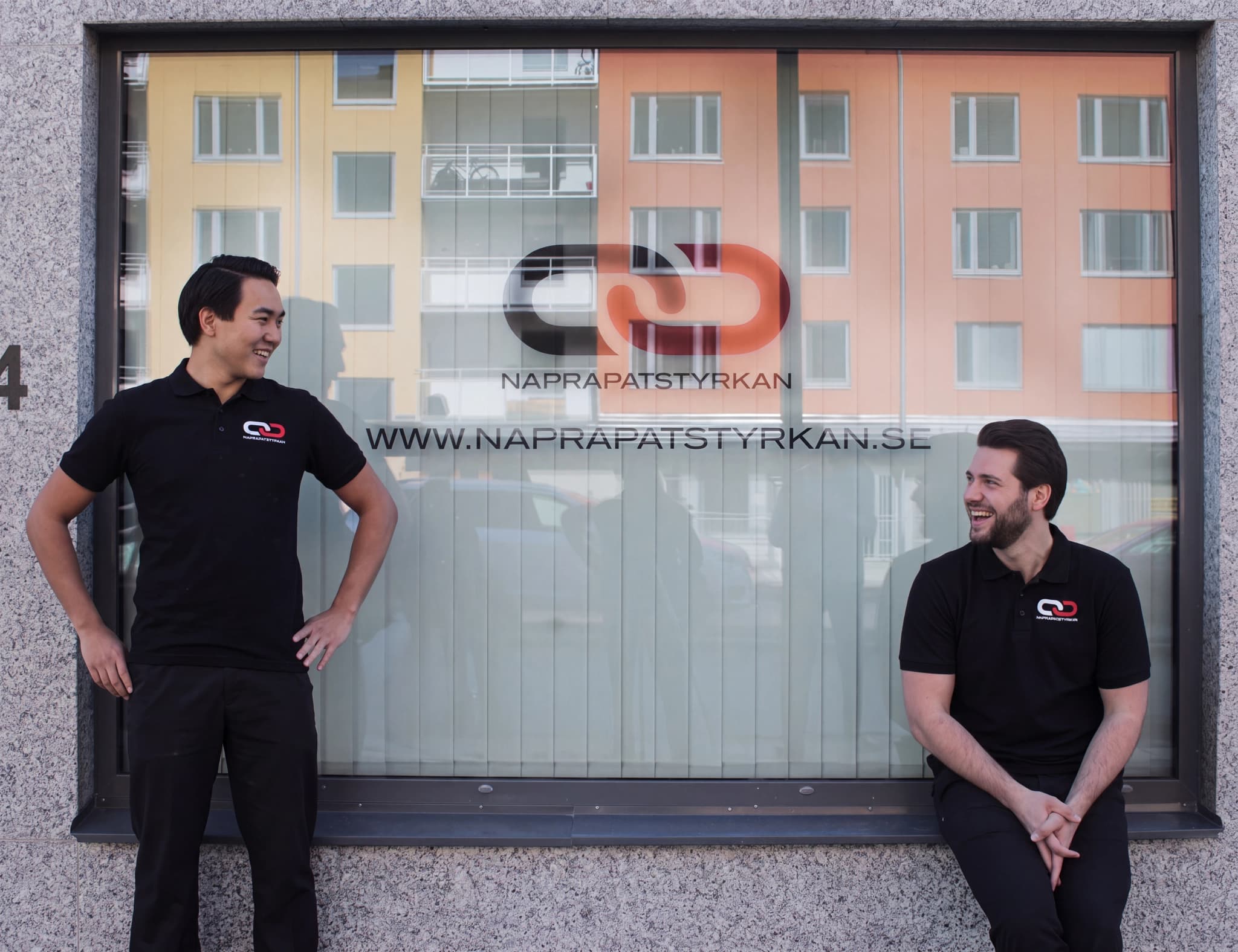 Två leende manliga anställda framför ett fönster med företagets logotyp.