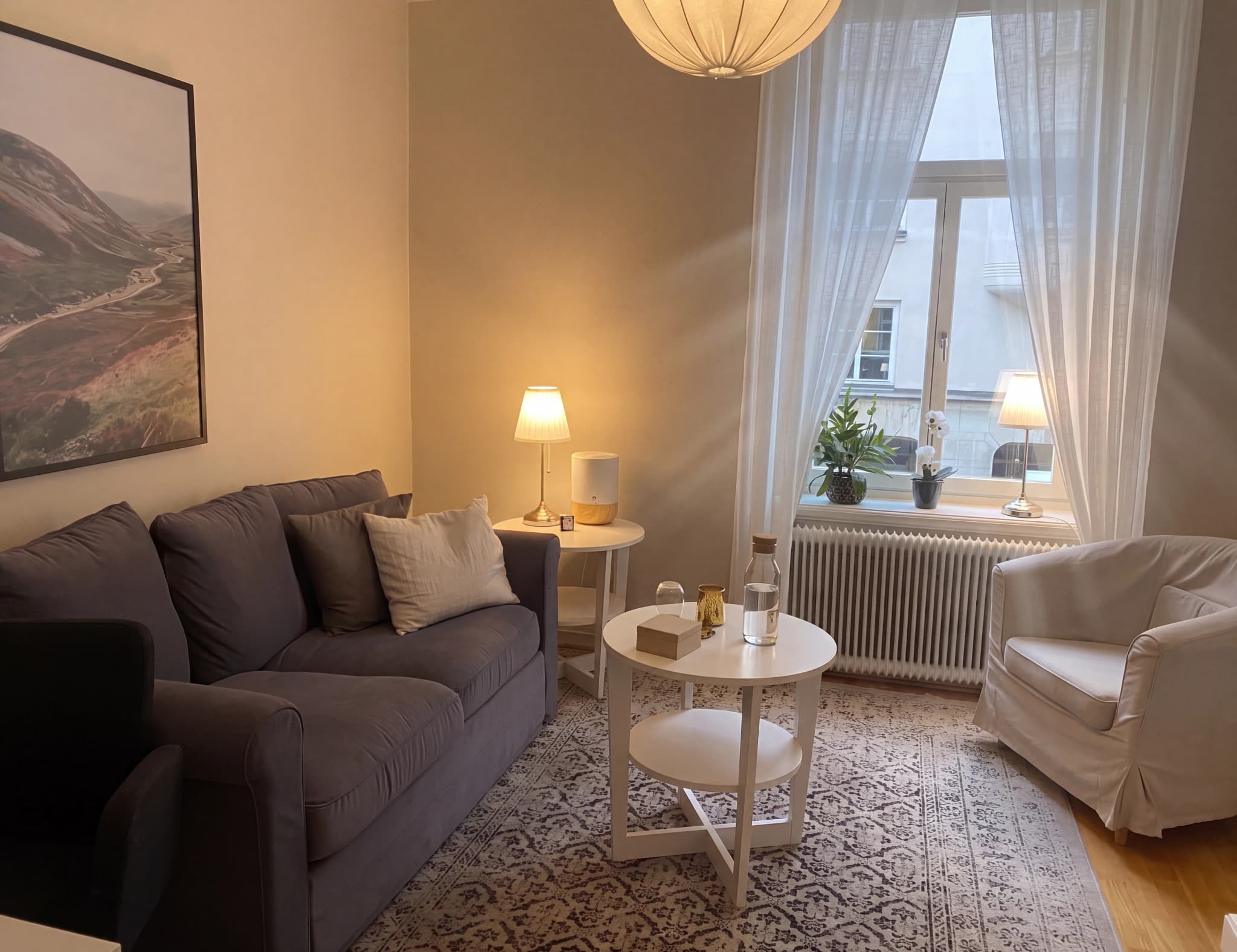 Ett mysigt terapirum med en mörkgrå soffa, en vit fåtölj, ett runt soffbord och fönster med vita gardiner.