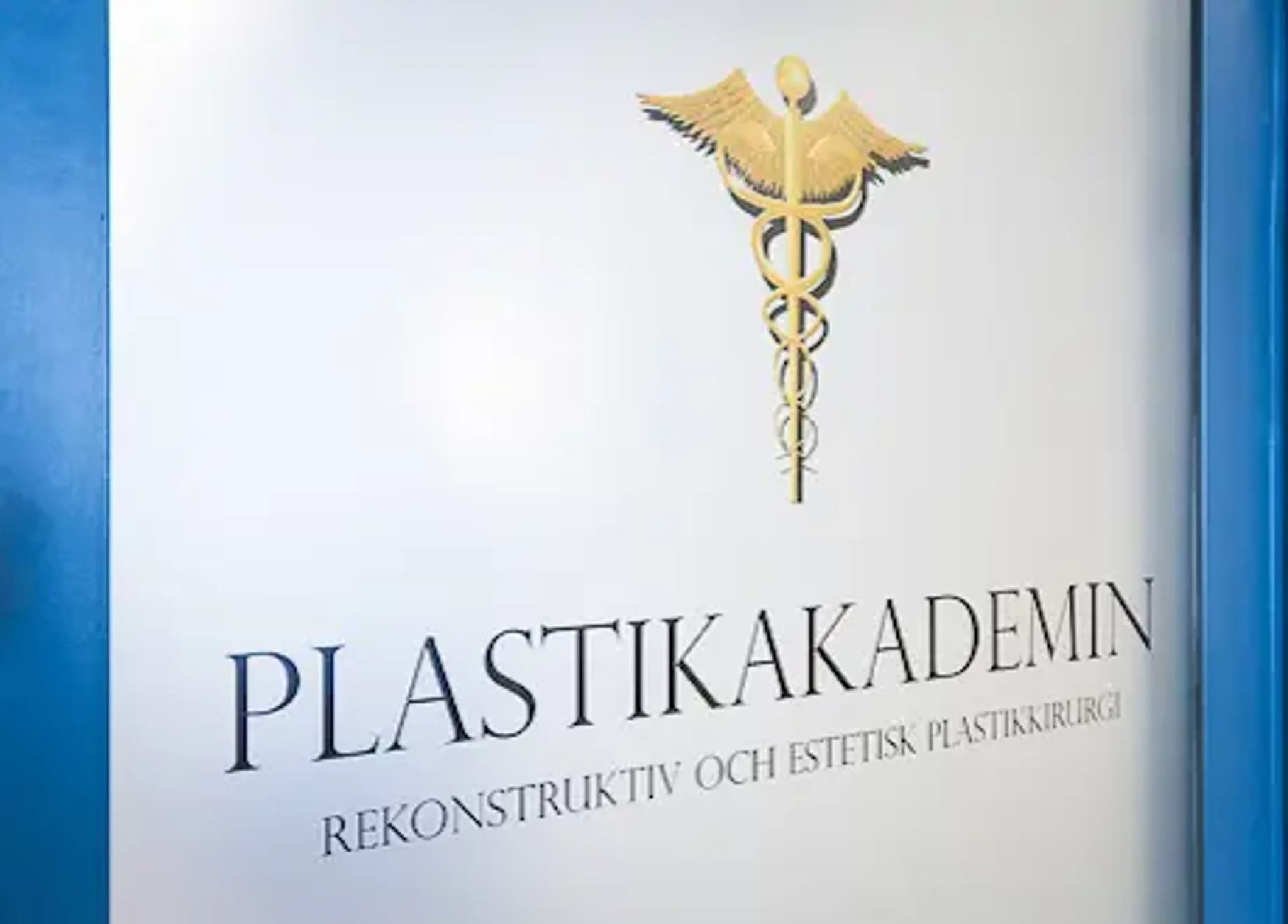 Plastikakademin-logotyp med en gyllene Caduceus-symbol ovanför texten "PLASTIKAKADEMIN" och "REKONSTRUKTIV OCH ESTETISK PLASTIKKIRURGI".