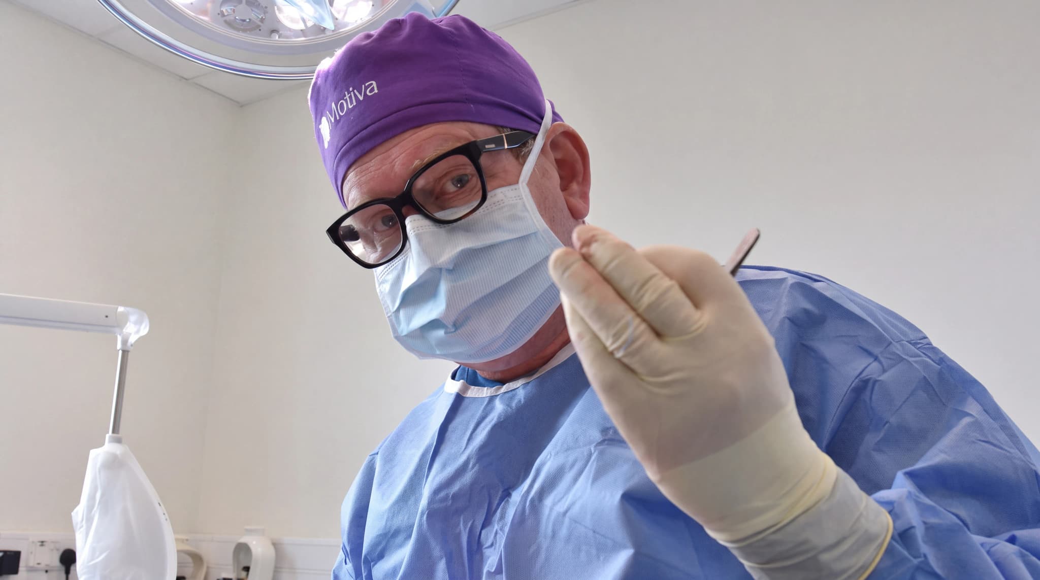 Kirurg med munskydd och glasögon tittar mot kameran och håller ett instrument i handen, klädd i blå operationskläder och lila mössa.