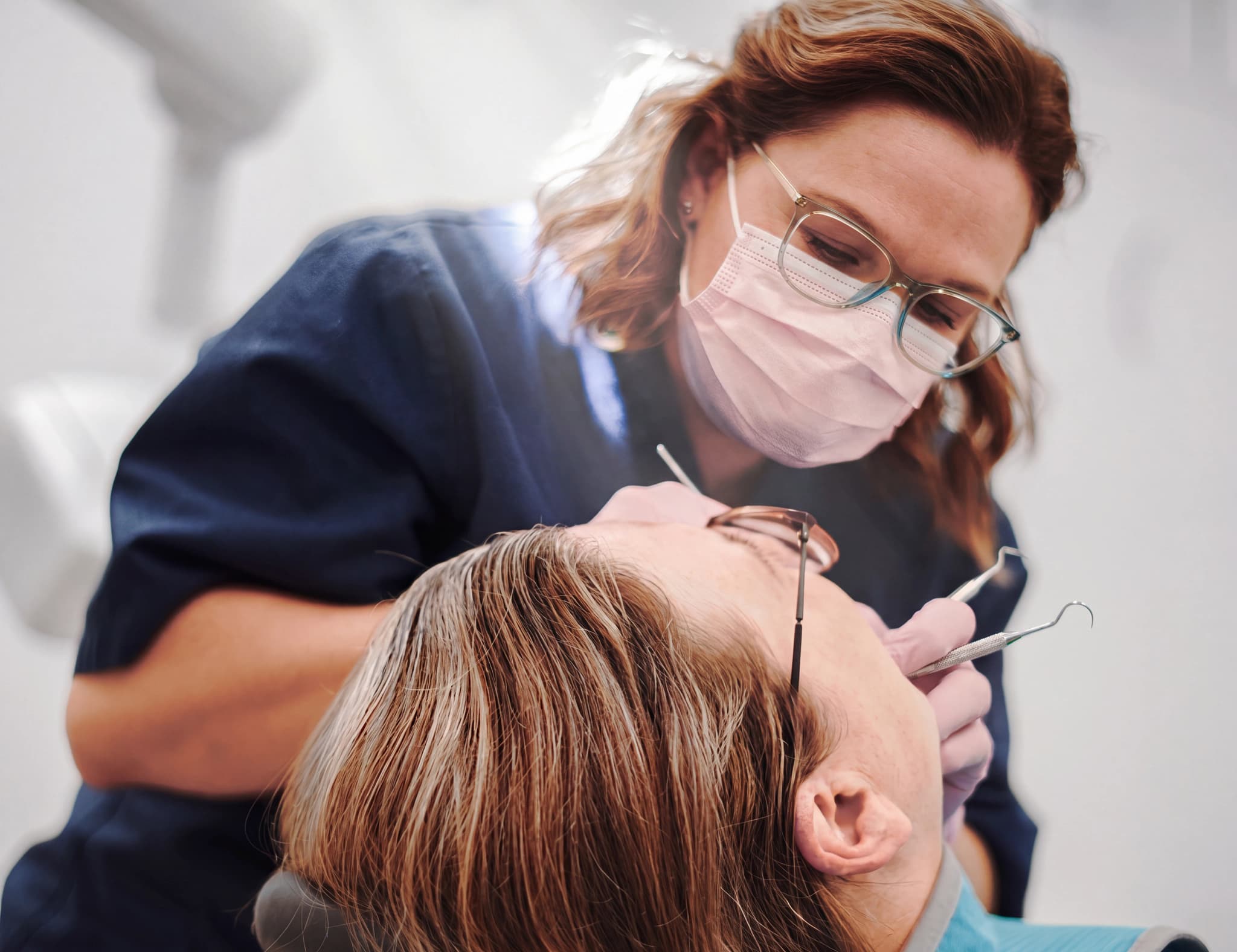 Tandläkare med munskydd och glasögon undersöker en patients mun med ett instrument.