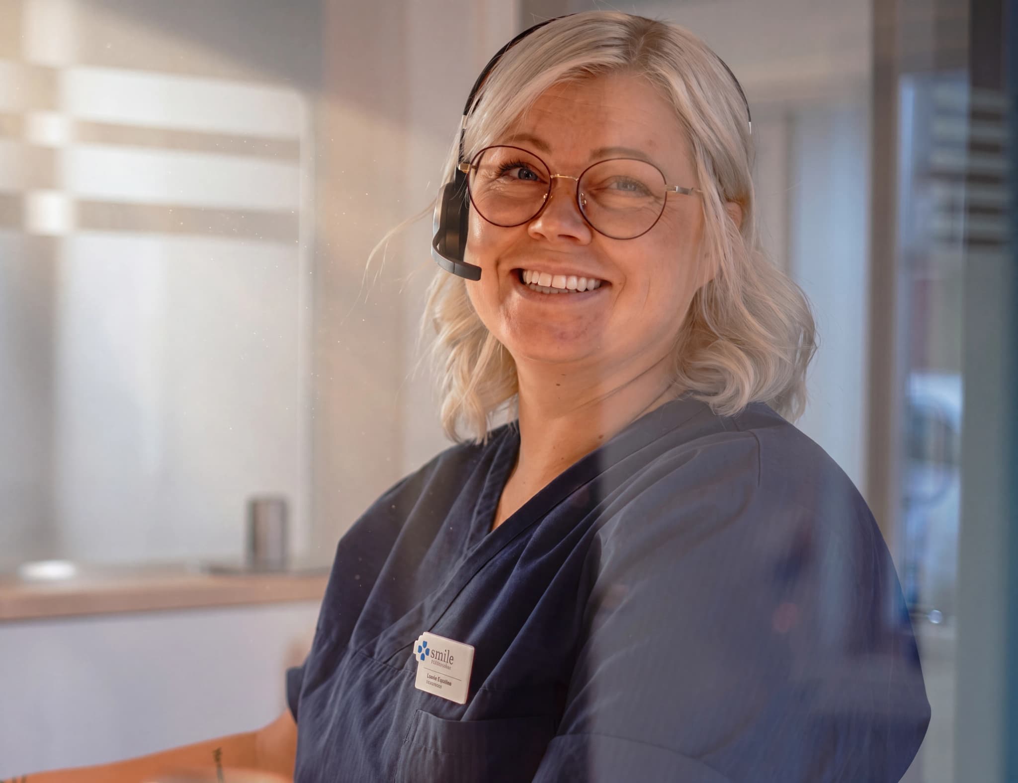 En kvinna med glasögon och headset ler när hon sitter vid ett skrivbord.