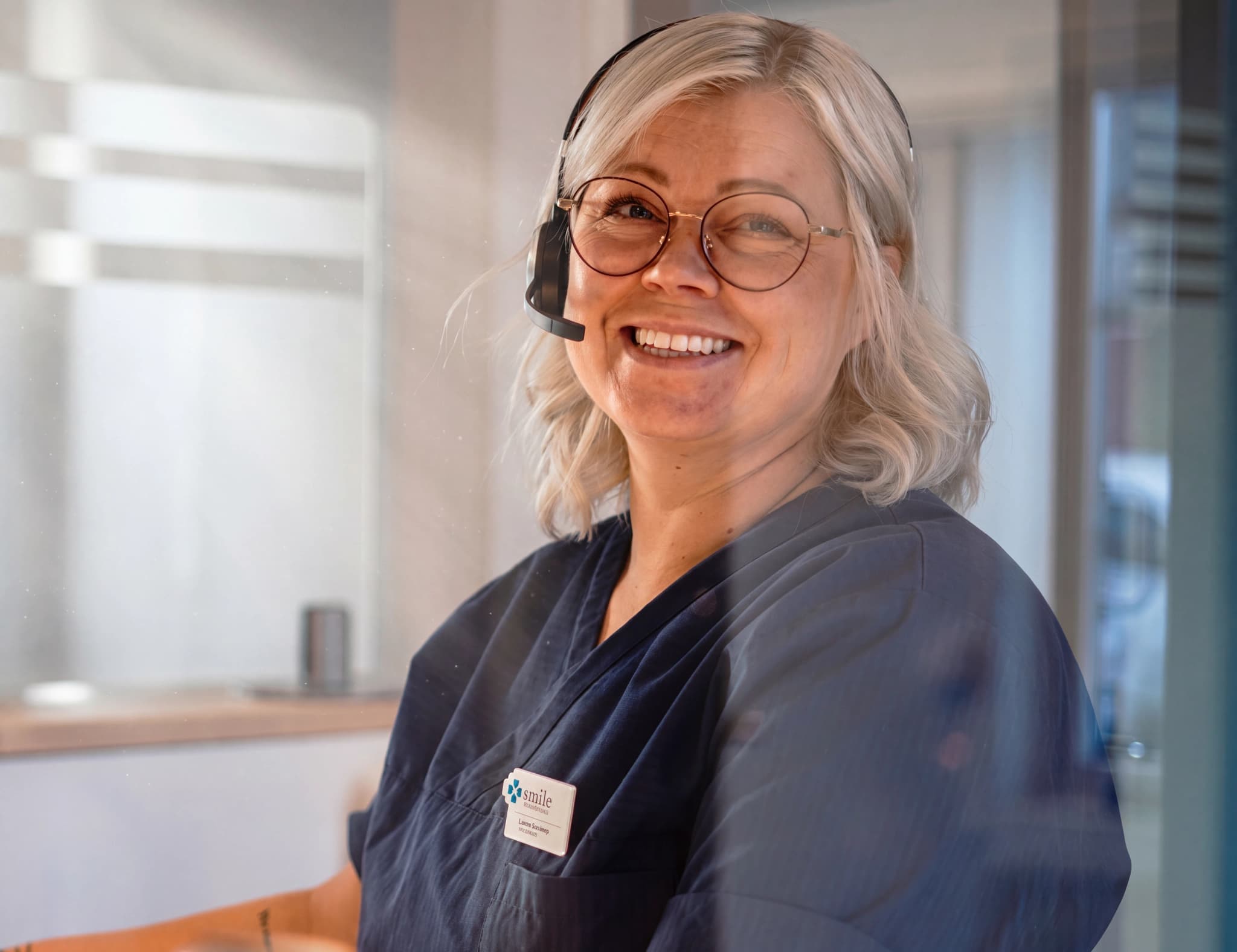En kvinna med glasögon och headset ler mot kameran, hon bär en mörkblå klädsel med ett namnskylt