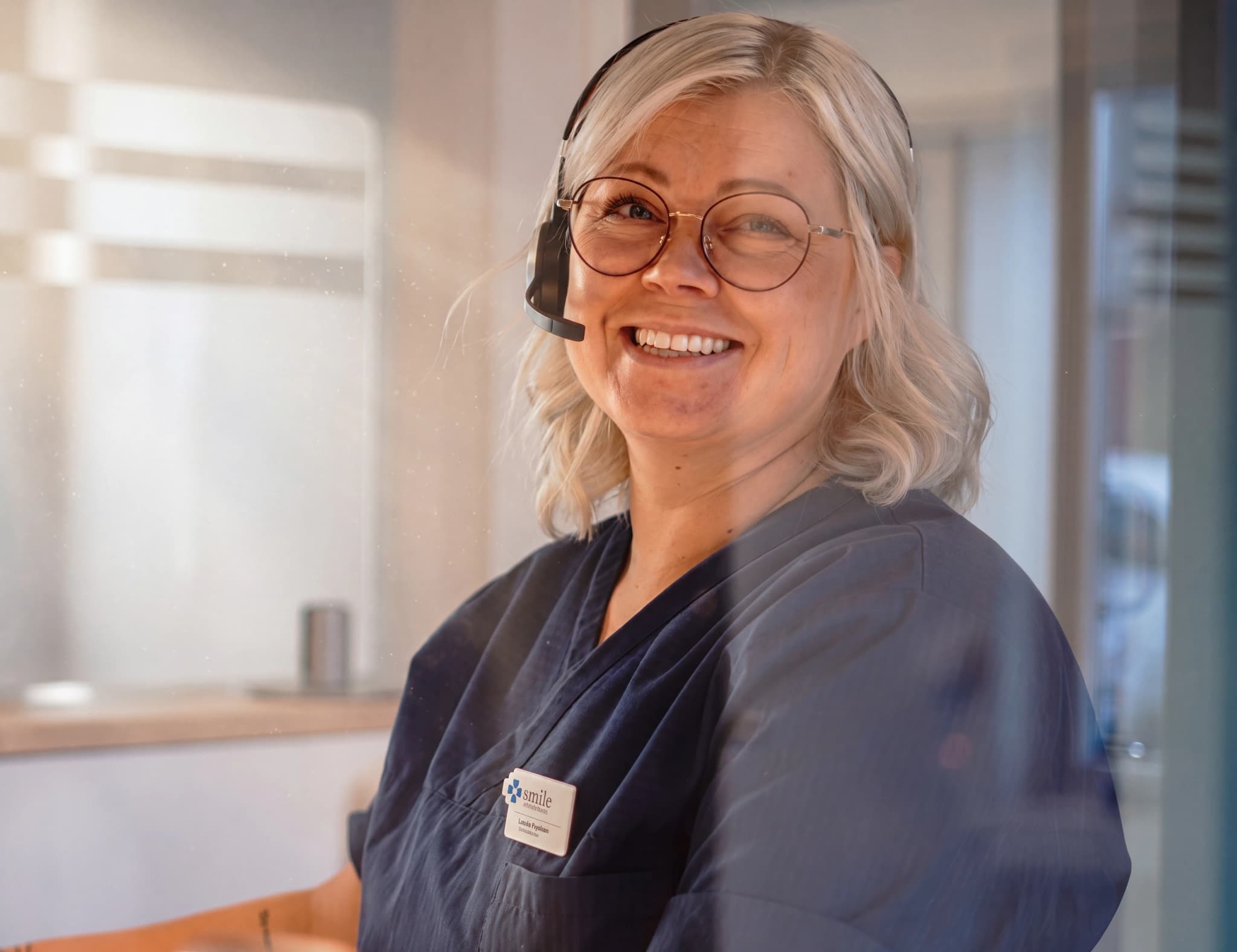 En kvinna med glasögon och headset ler mot kameran vid en reception i en klinik.