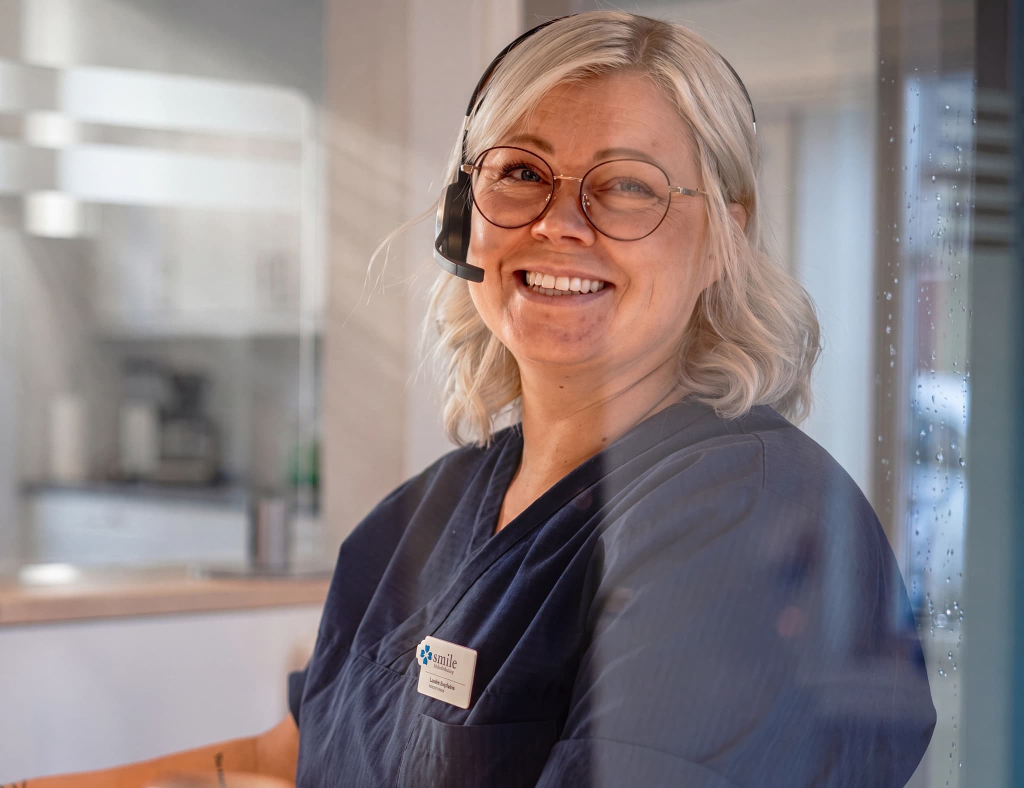 En kvinna med glasögon och headset ler mot kameran, iklädd en mörkblå arbetsklädsel med en namnskylt.