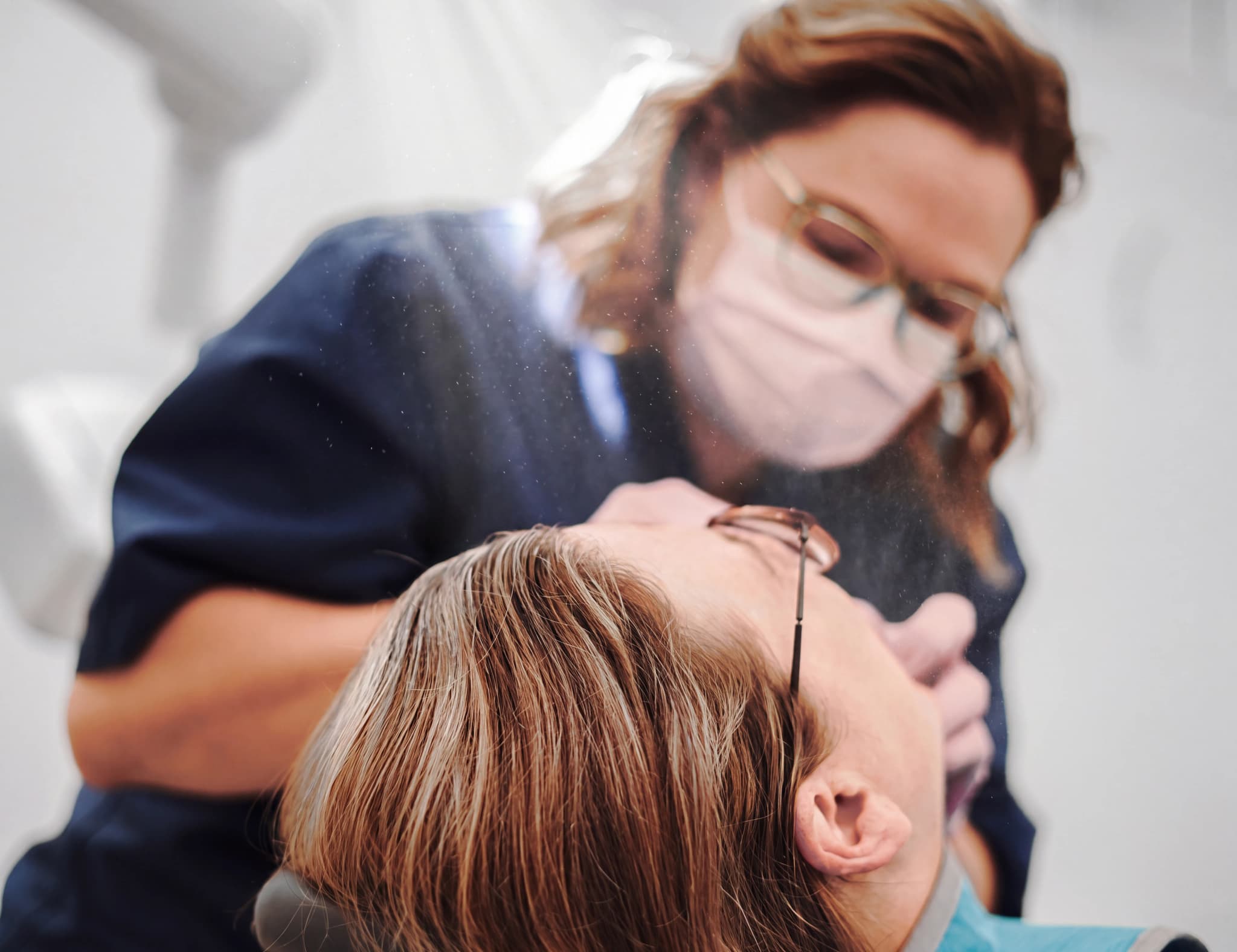 Tandläkare i munskydd och glasögon undersöker en patients mun med instrument.