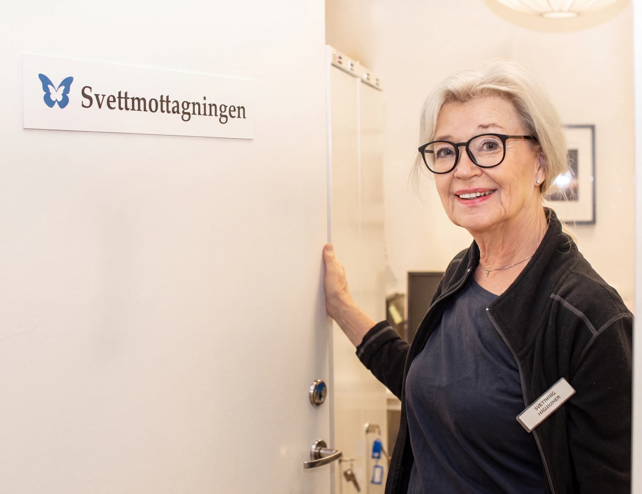 En kvinna med glasögon ler och står bredvid en dörr med en skylt som det står "Svettmottagningen" på.