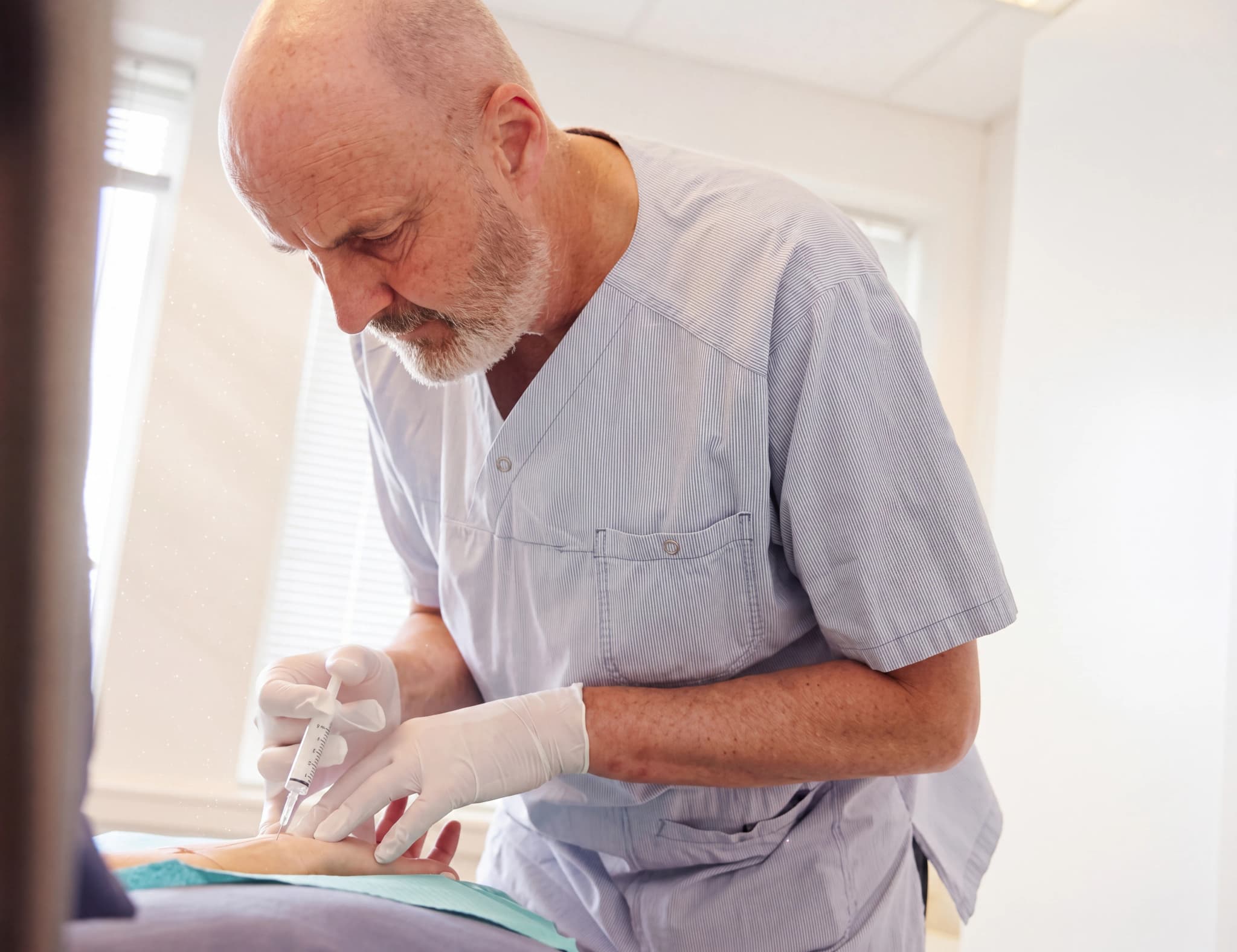 Läkare med skägg i ljusblå kläder med gummihandskar som ger en patient en injektion i armen.