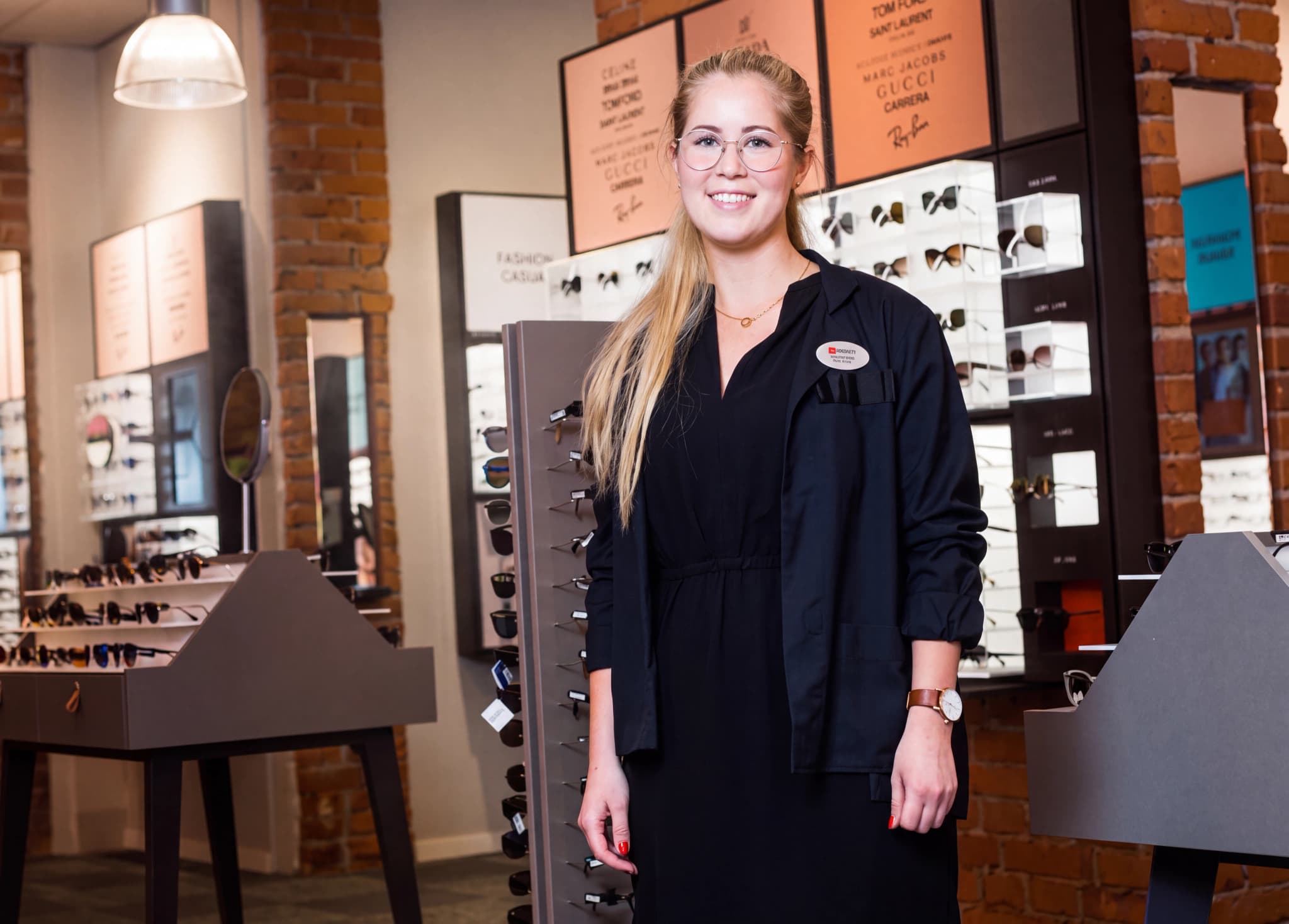 En leende optiker står framför en display med glasögon i en butik.