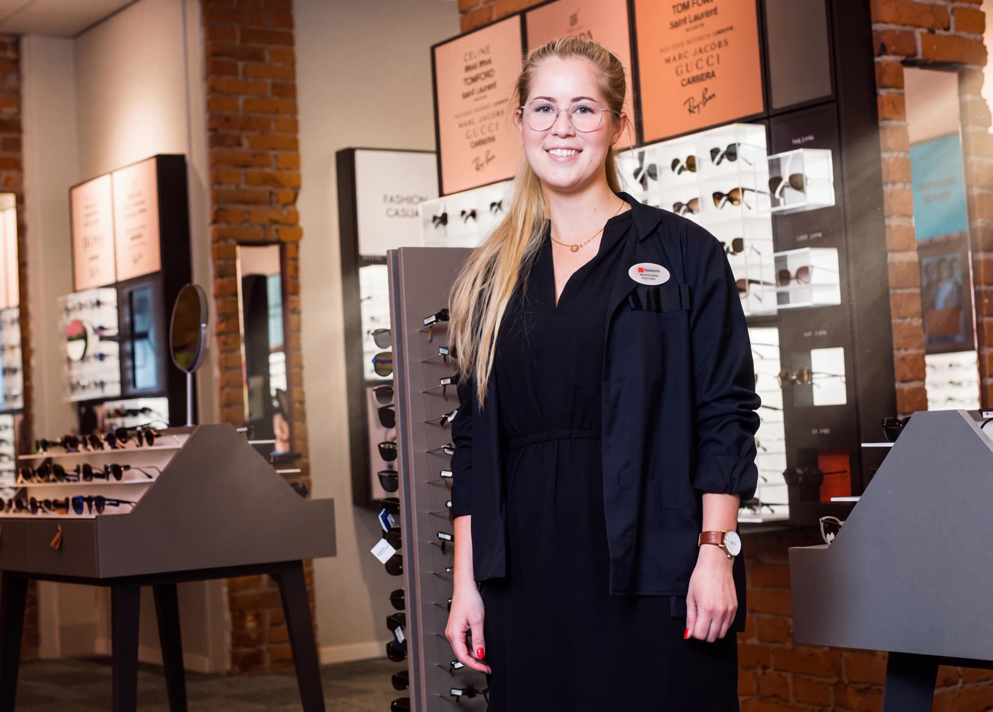 En kvinna med glasögon och en svart tröja med ett märke står i en optikerbutik och ler mot kameran.