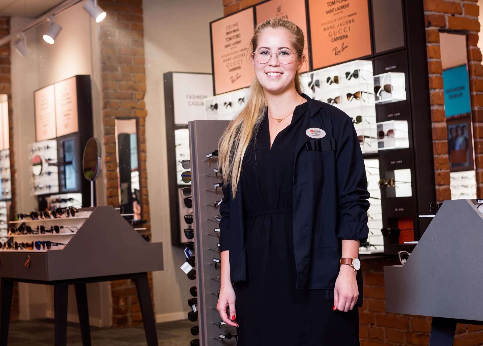 En ung kvinna med glasögon och en svart uniform står i en butik som säljer glasögon och solglasögon.