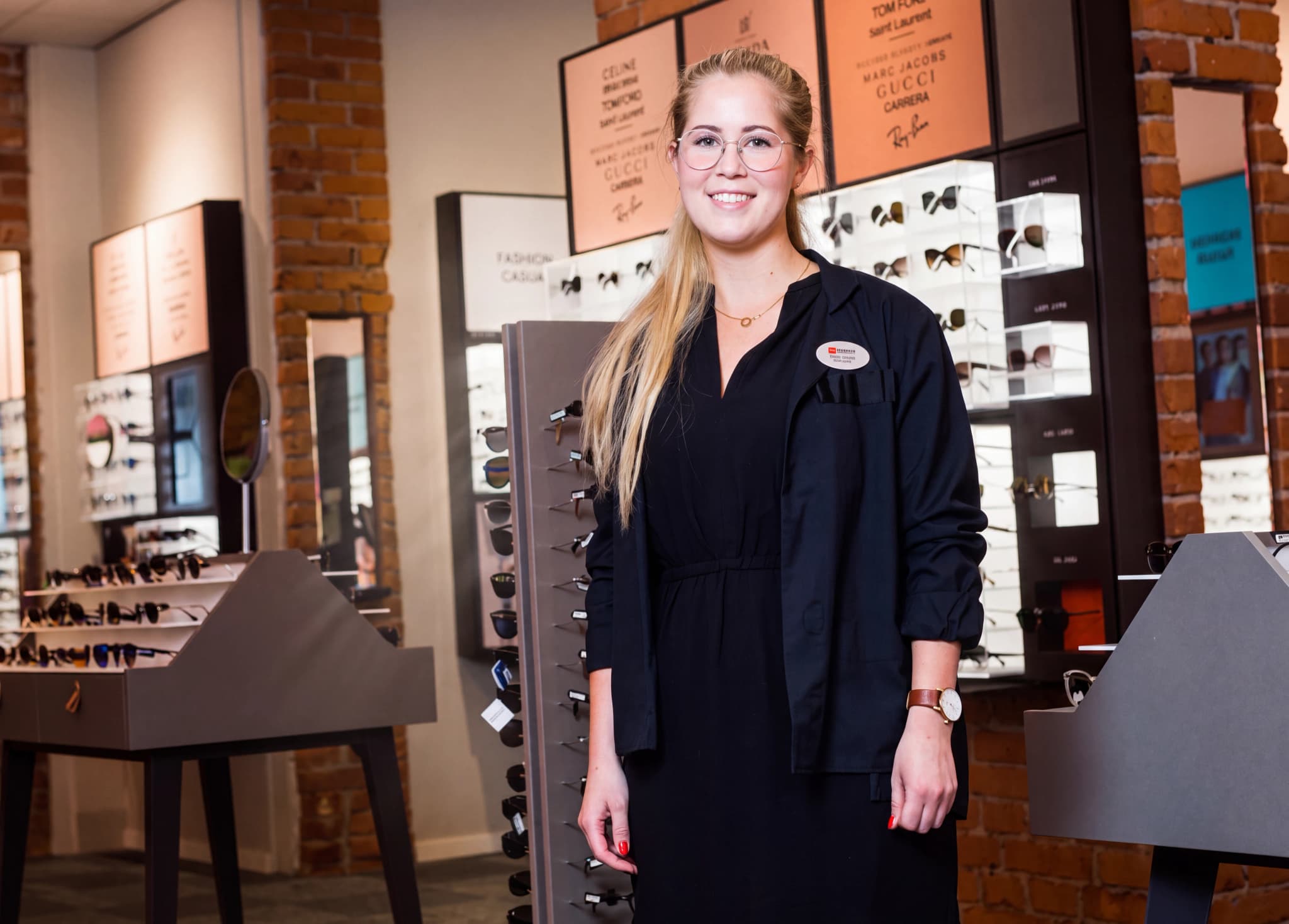 En leende optiker står i en butik med glasögon och solglasögon på display.