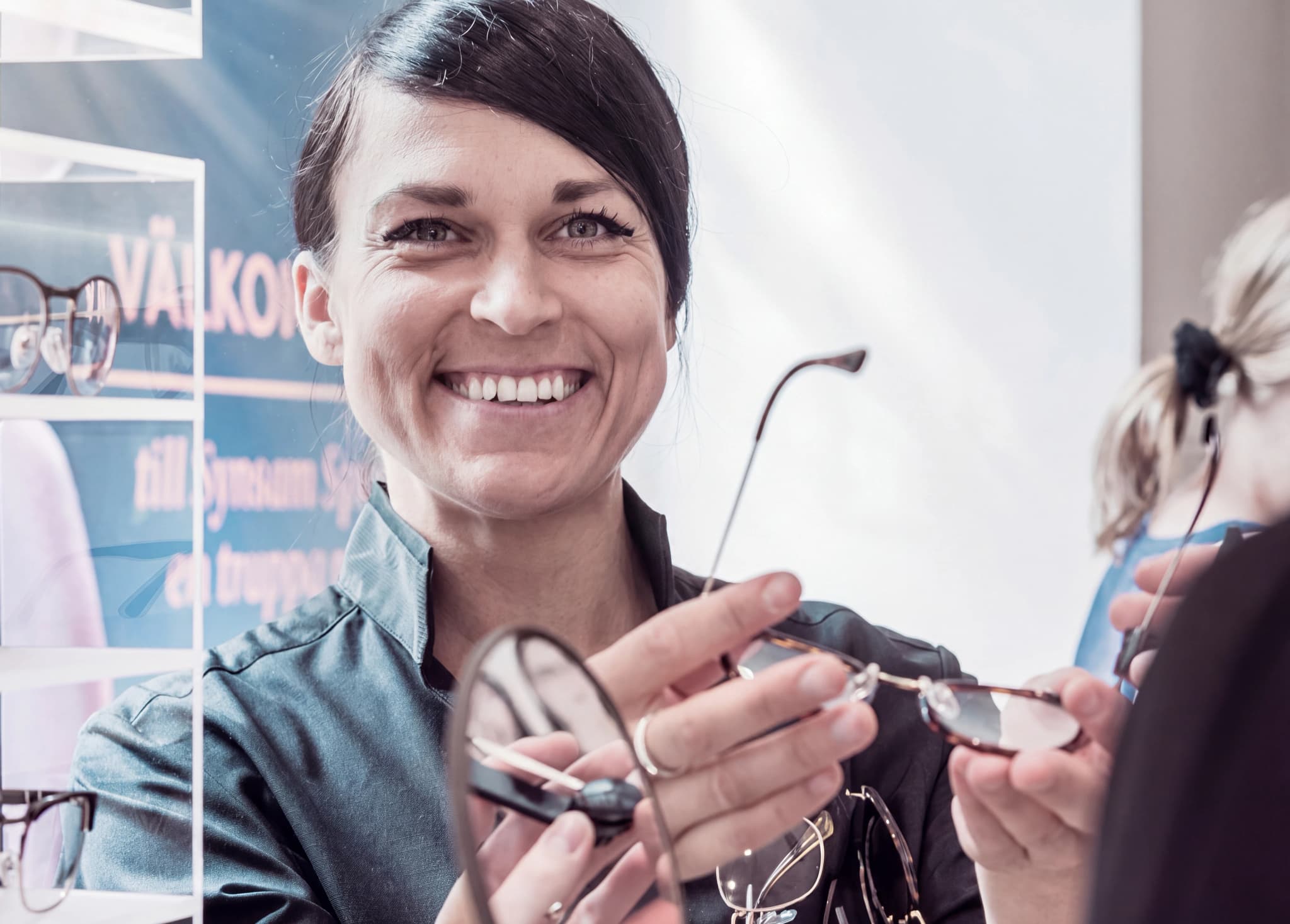 En kvinnlig optiker ler och håller upp ett par glasögon för en kund i en butik.