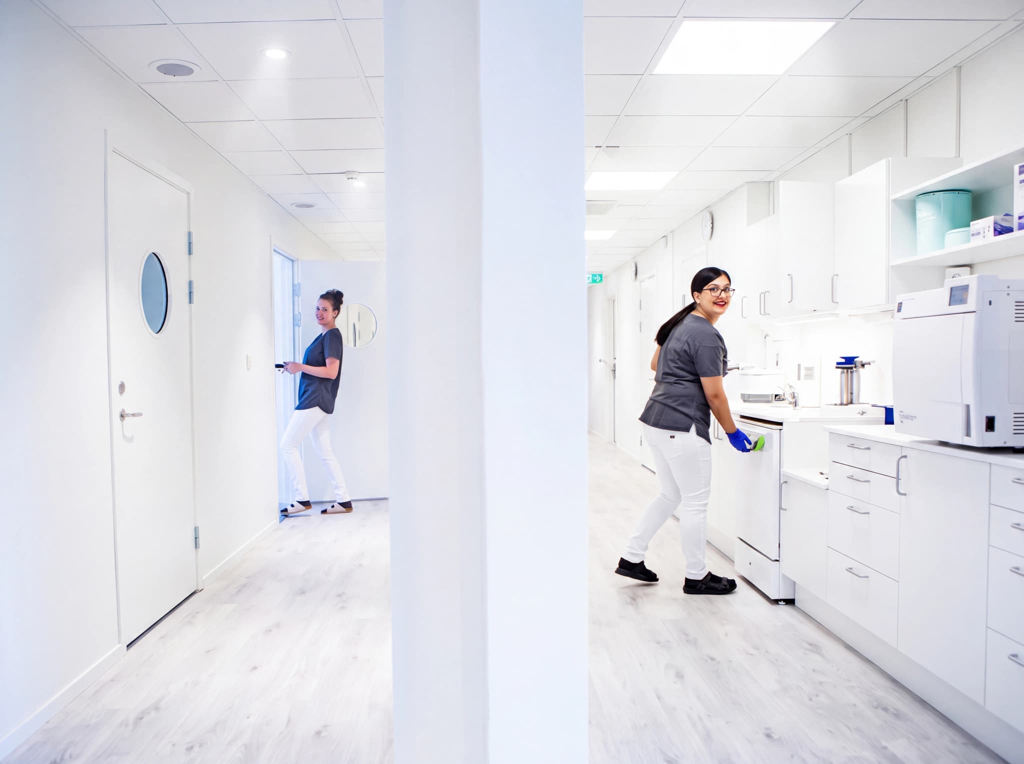 Två kvinnliga vårdpersonal arbetar i en ljus och modern klinikkorridor. Den ena står vid en dörr och den andra vid medicinsk utrustning.