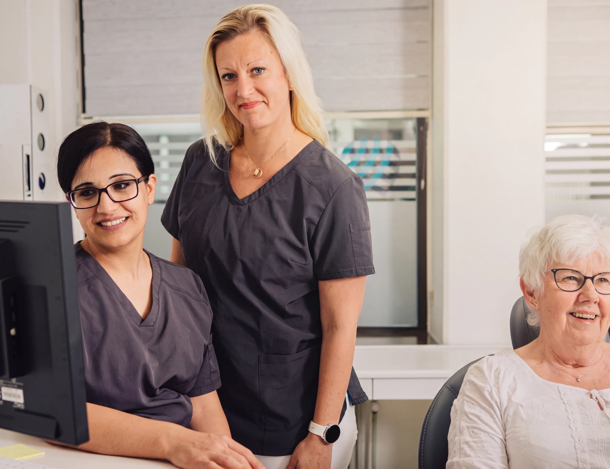 Två kvinnliga tandvårdspersonal tittar på en datorskärm, medan en äldre patient sitter i en tandläkarstol.