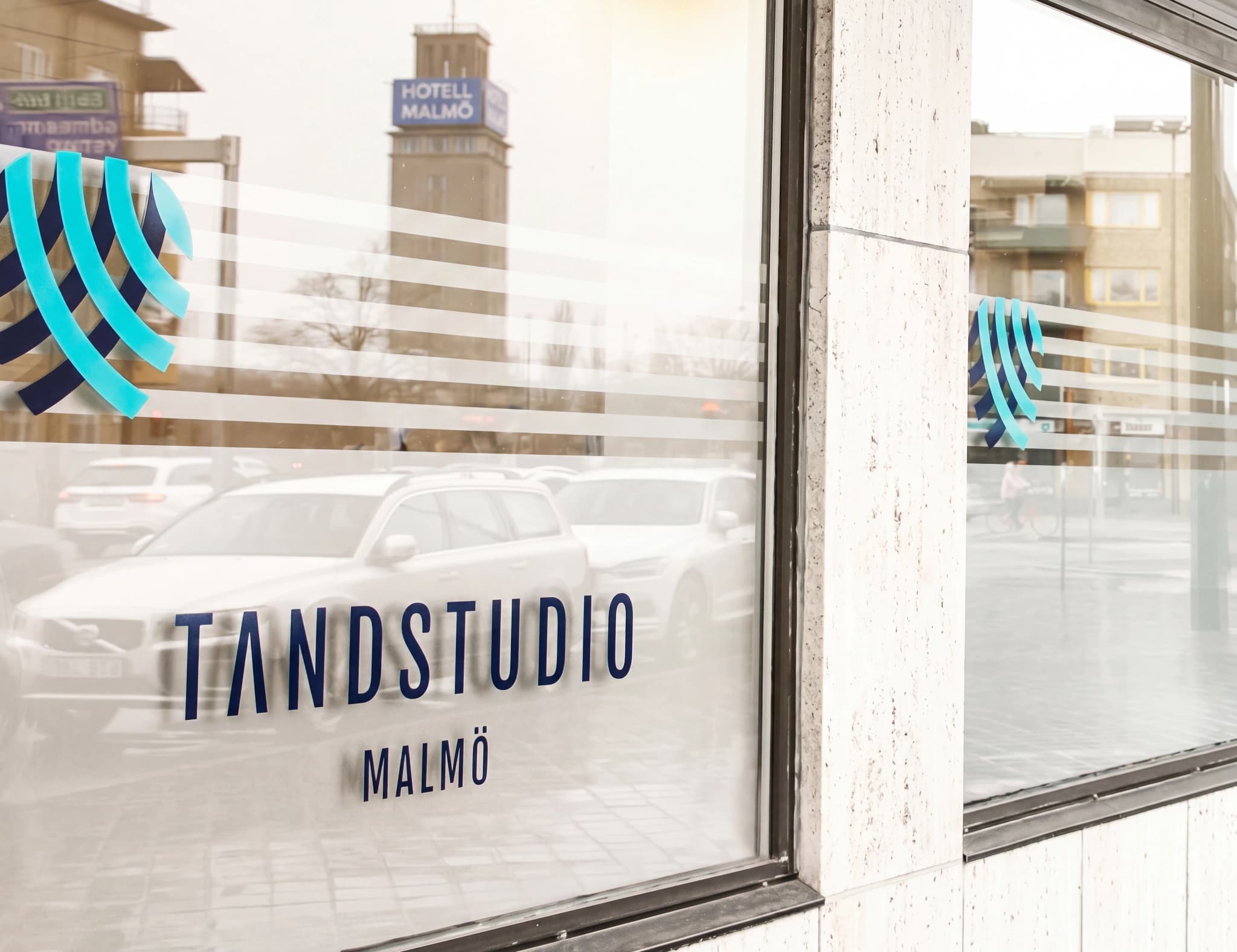 Texten Tandstudio Malmö och en logotyp med blå linjer syns på ett fönster med bilar och en byggnad i bakgrunden.