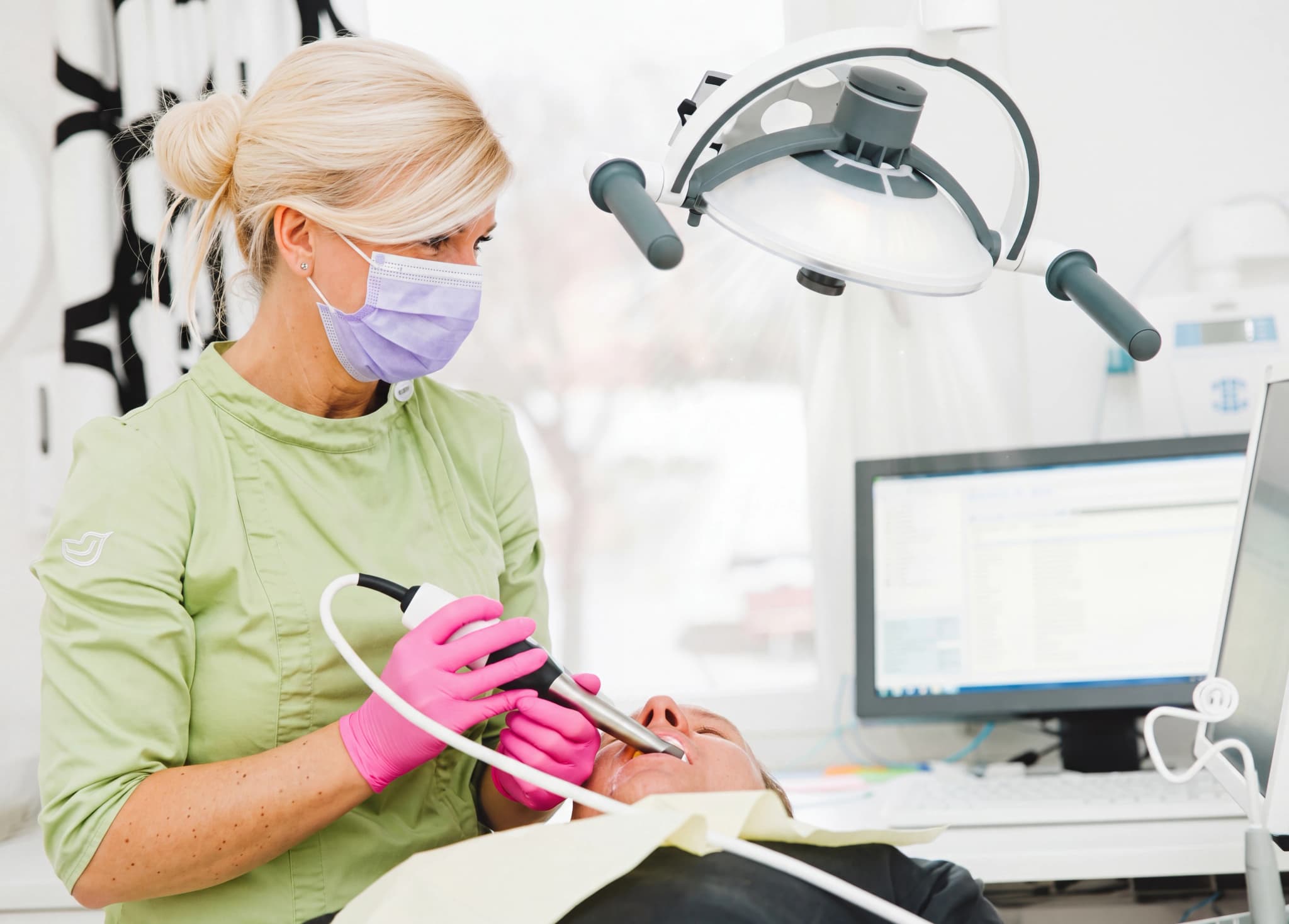 Tandläkare med munskydd och rosa handskar använder en dentalborr för att rengöra en patients tänder.