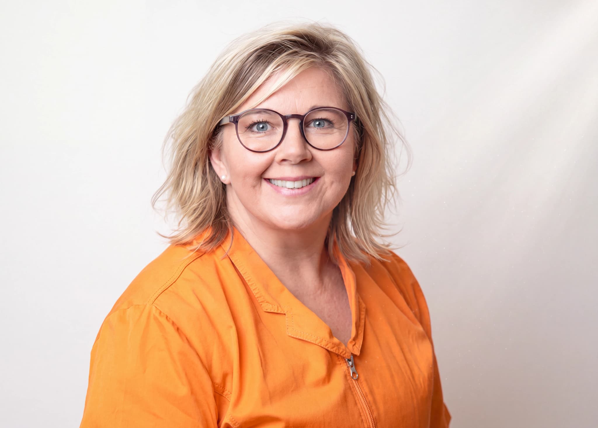 En kvinna med blont hår och glasögon ler in i kameran, klädd i orange arbetskläder.