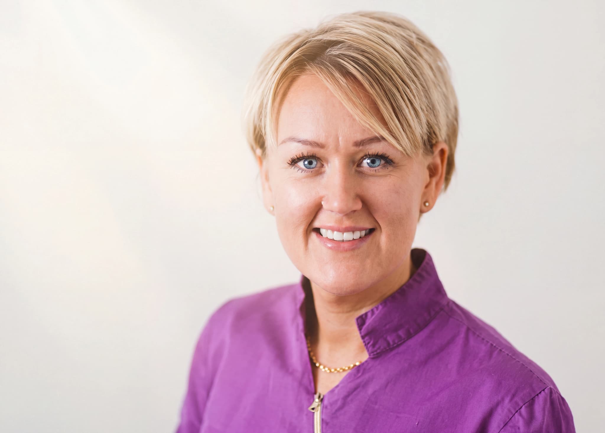Kvinna med blont hår och blå ögon ler glatt i en lila uniform.