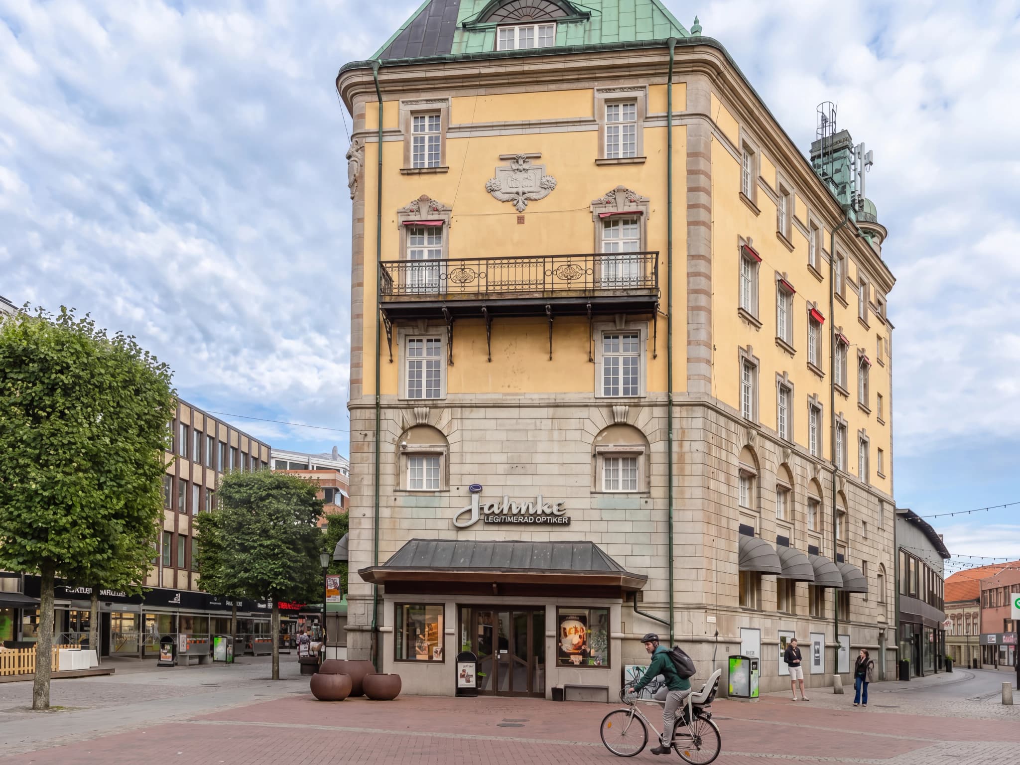 Byggnadens gula fasad med fönster och en balkong, med en optikerbutik på bottenvåningen.