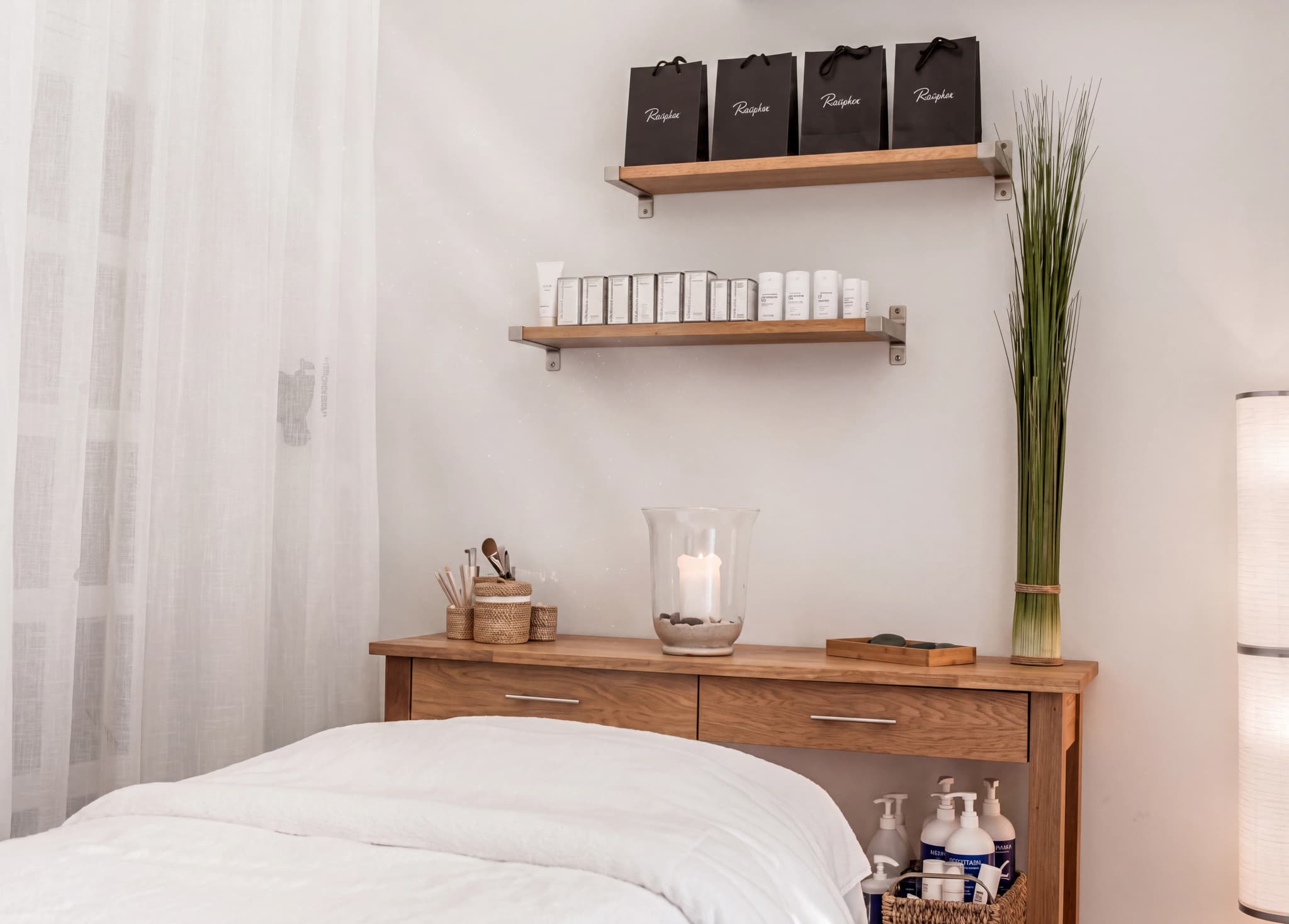 Spa-behandlingsrum med ett uppbäddat massagebord, hyllor med produkter, en vas med högt gräs och en lampa.