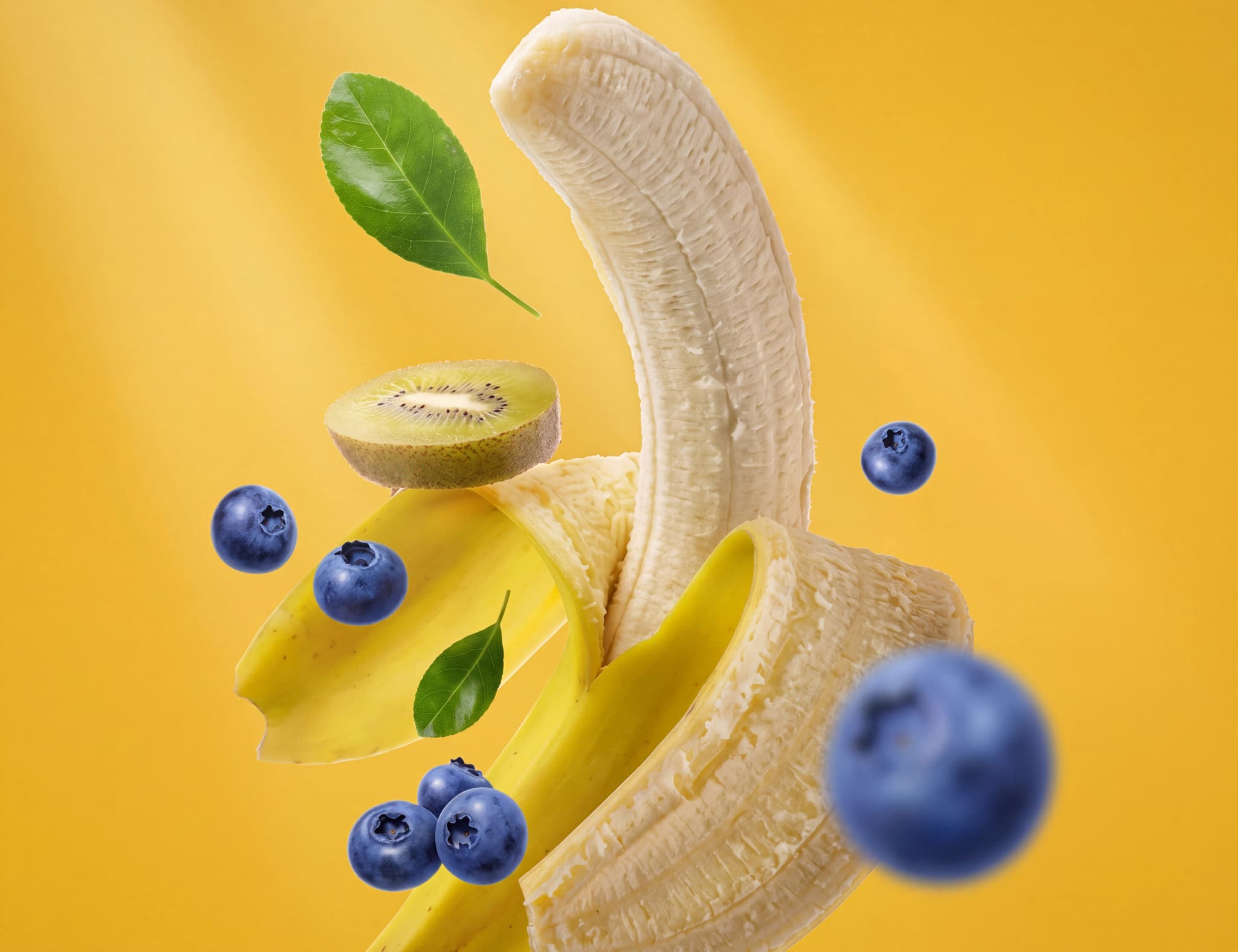 En skapad bild med en delvis skalad banan, en skiva kiwi, blåbär och gröna blad mot en gul bakgrund.