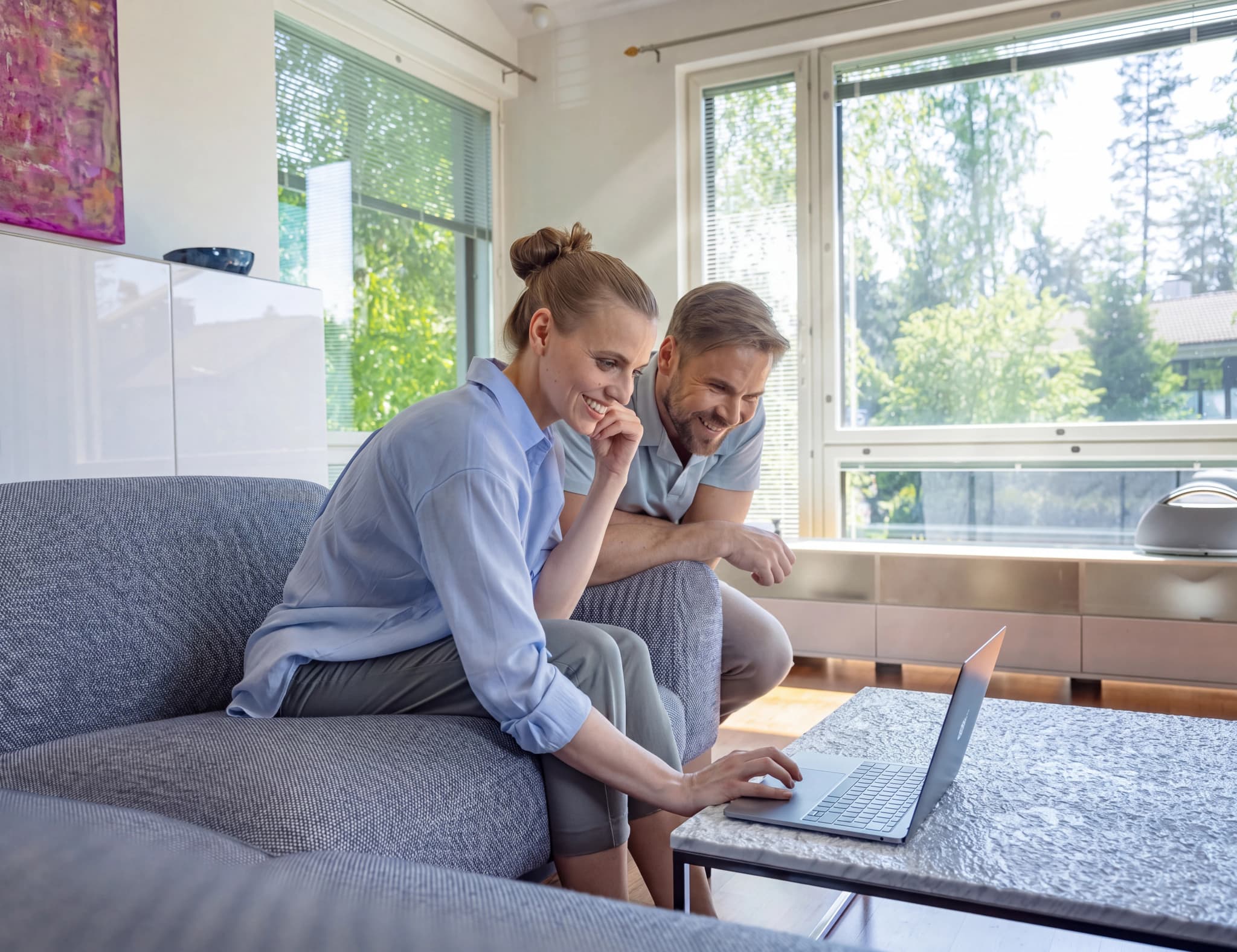 Ett leende par sitter på en soffa och använder en laptop tillsammans, med ett fönster med grönska i bakgrunden.