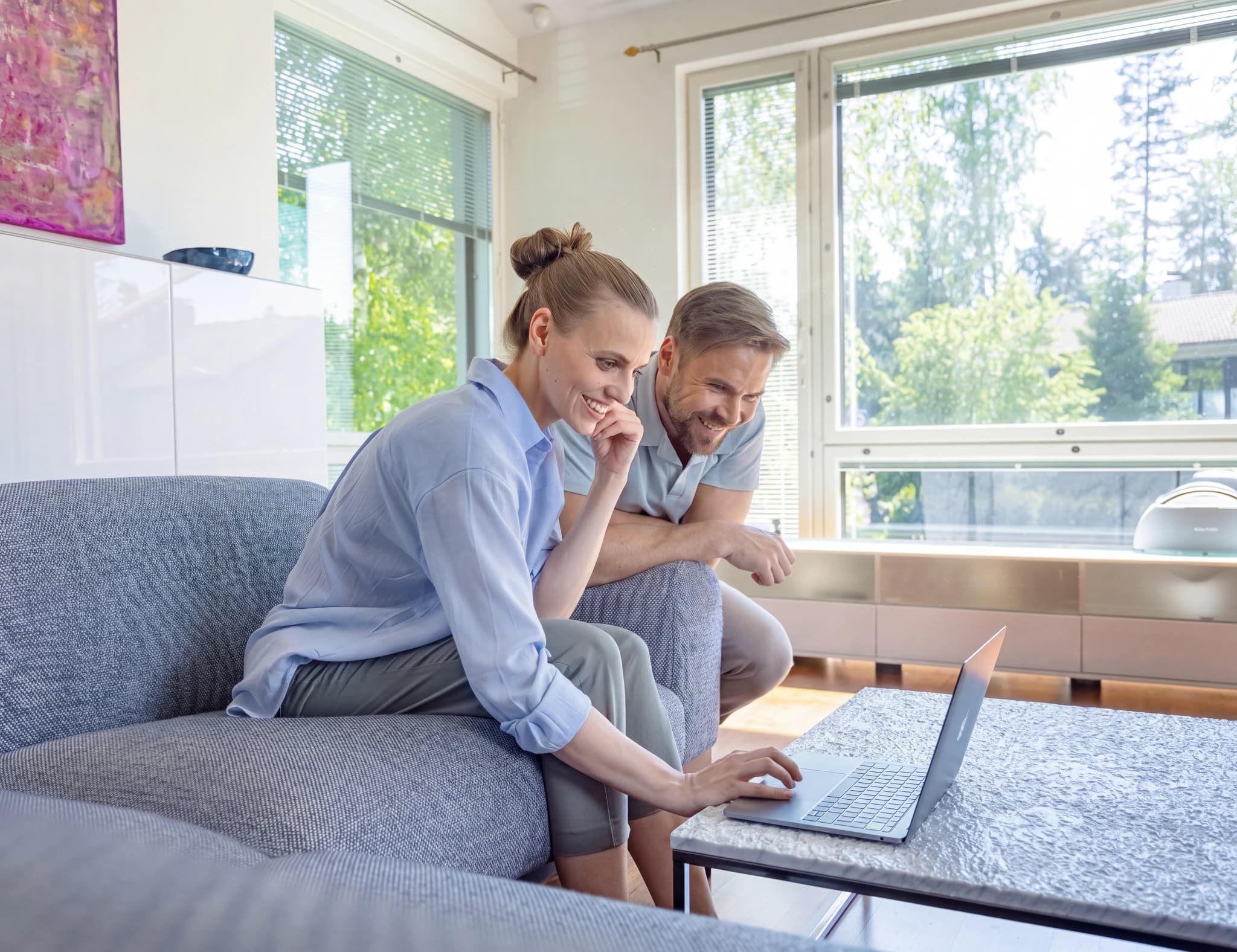 Ett par tittar och ler mot en laptop som ligger på ett soffbord medan de sitter på en soffa