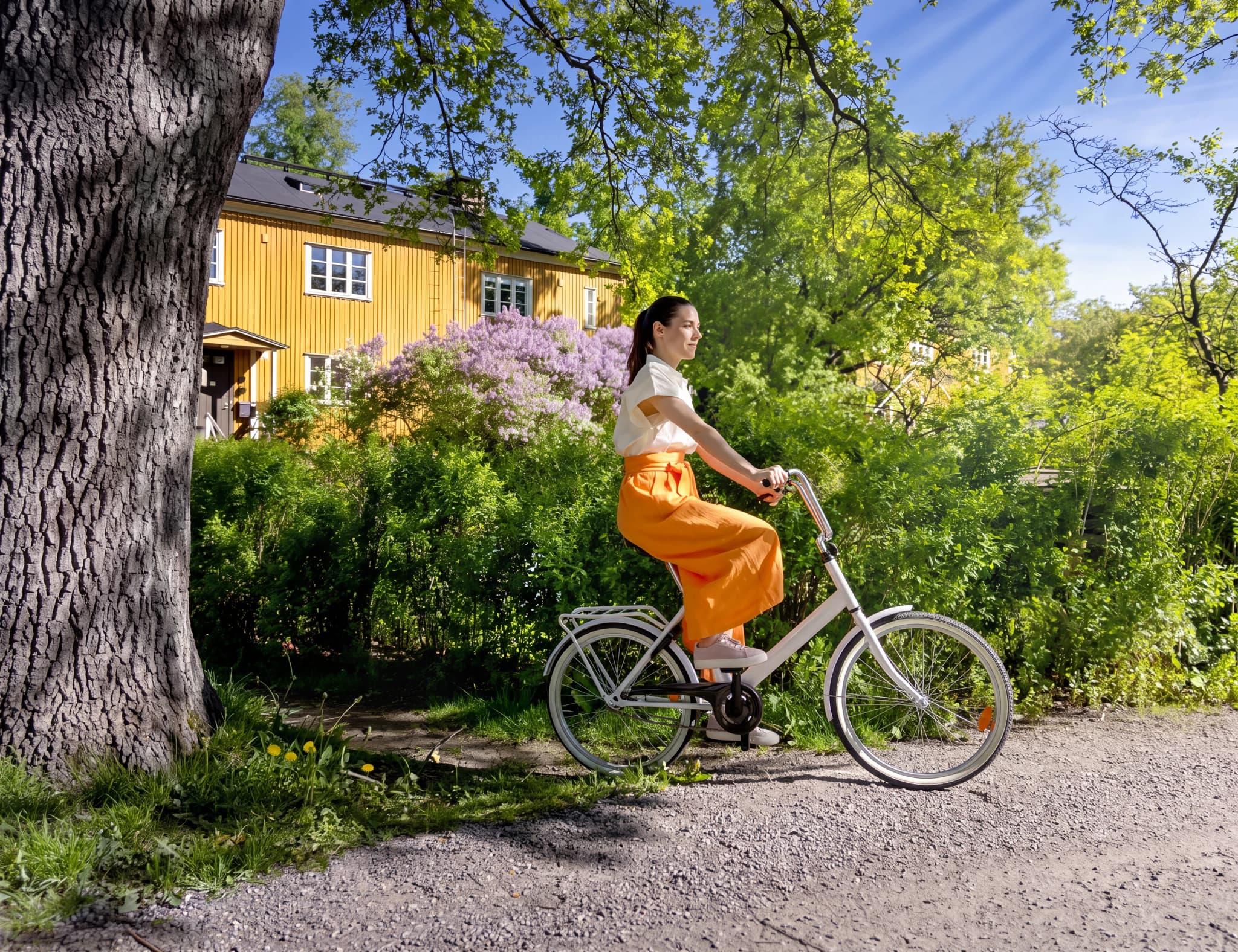 Kvinna cyklar på en grusväg nära ett gult hus med lila blommor.