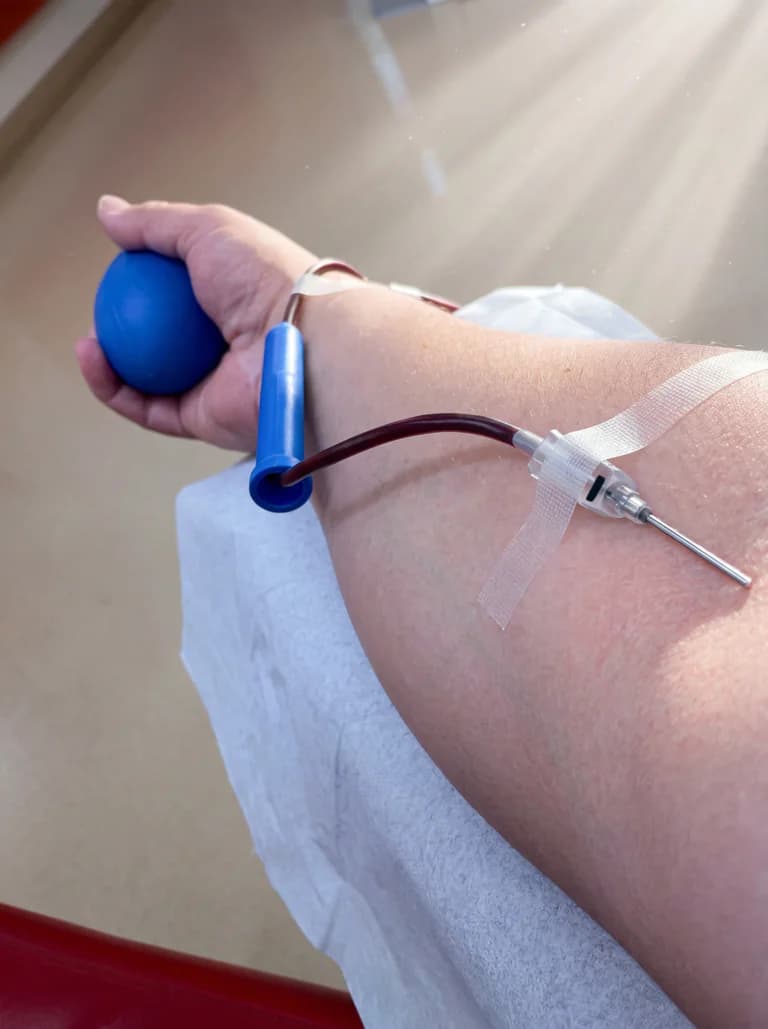 En person som donerar blod, håller en blå stressboll i handen när nålen är instucken i armen.