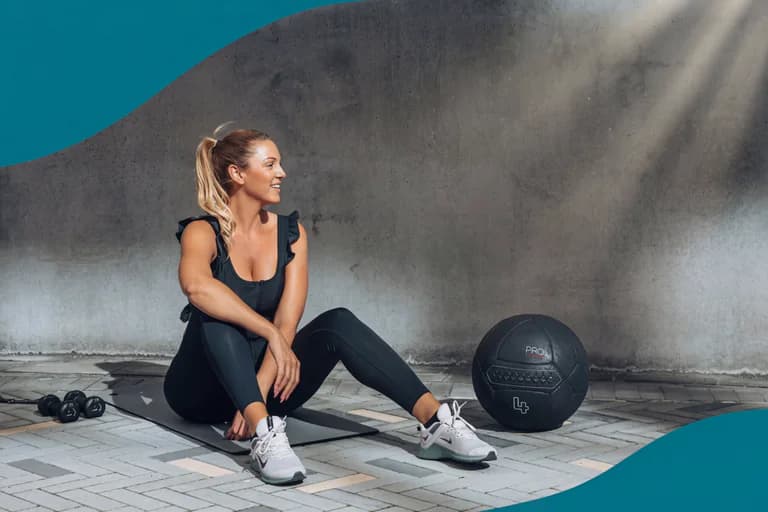 En kvinna i träningskläder sitter på en yogamatta bredvid en medicinboll och ett par hantlar.