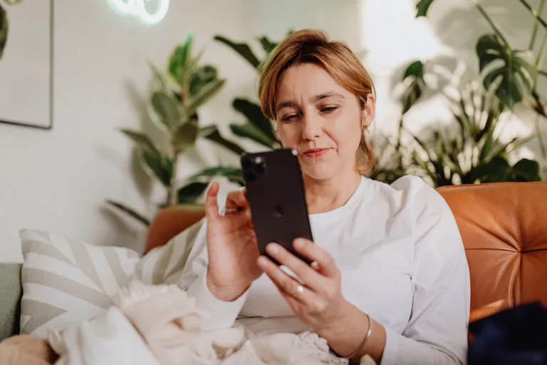 En kvinna i 40-årsåldern sitter på en soffa och använder sin smartphone. Bakgrunden visar inomhusväxter och en brun lädersoffa.