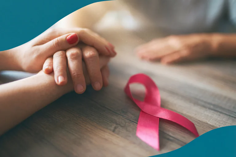 Två par händer som håller varandra, med en rosa banderoll för bröstcancer awareness på ett träbord.