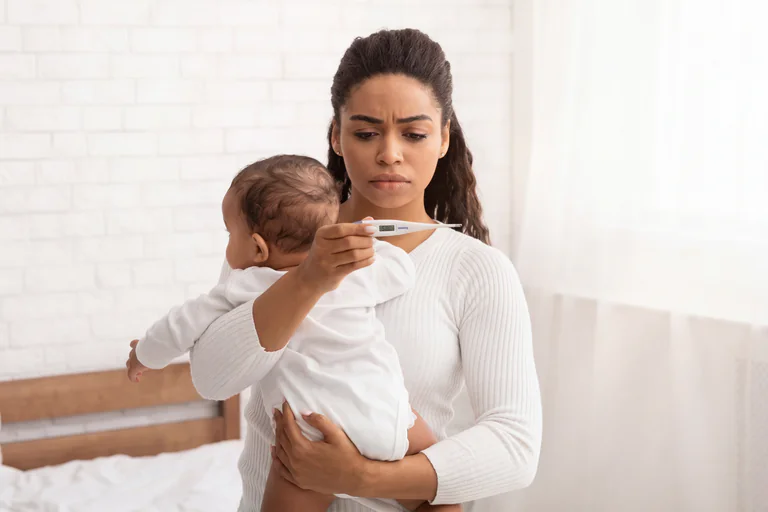 En orolig mamma håller ett spädbarn och kontrollerar dess temperatur med en termometer i ett sovrum.