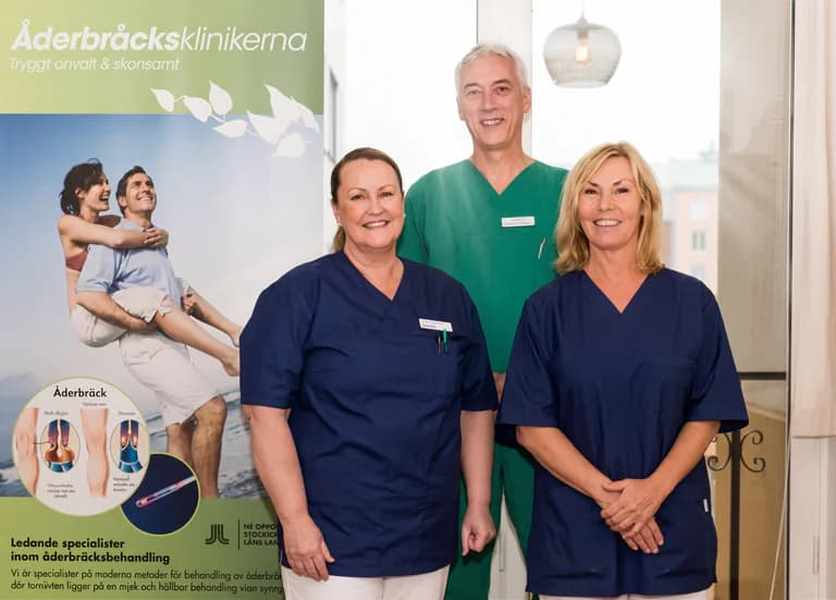 Tre leende vårdpersonal står framför en reklamaffisch för åderbråcksbehandling.