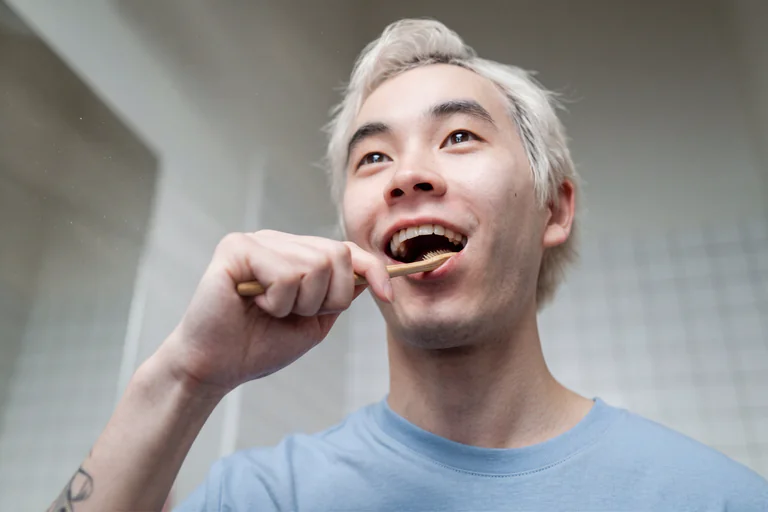En ung man med blont hår borstar tänderna med en bambutandborste i ett badrum.