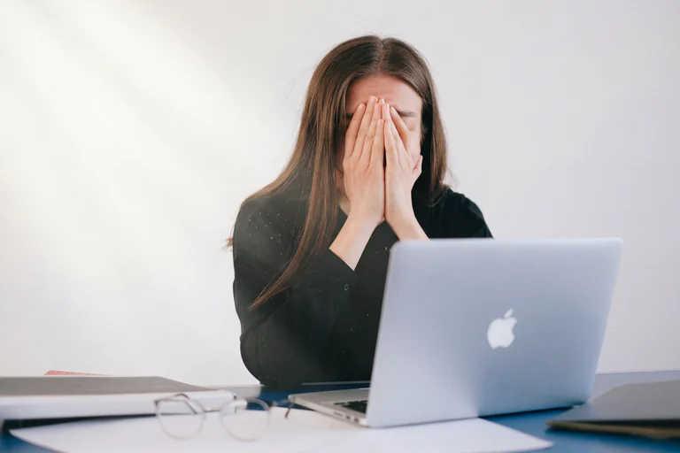 En kvinna med brunt hår sitter vid ett skrivbord med en bärbar dator framför sig och håller händerna över ansiktet, som om hon är upprörd eller trött. Ett par glasögon ligger på skrivbordet.